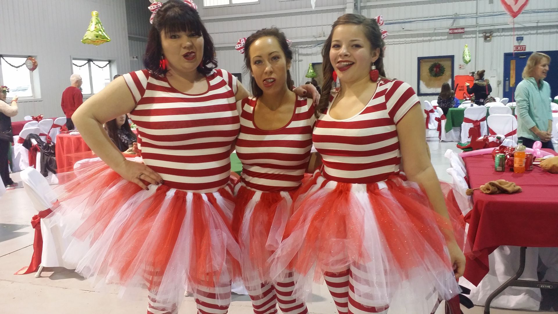 Three women in red-and-white striped outfits and tulle tutus pose at a festive indoor event.