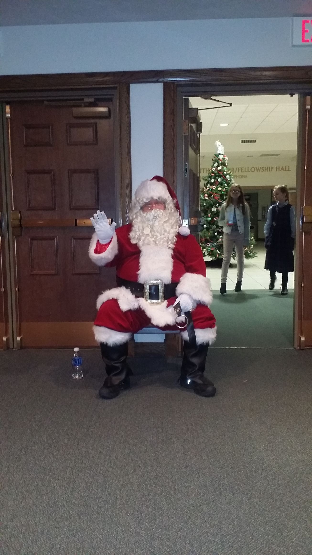 Santa Claus sitting and waving in a doorway, with a lit Christmas tree inside the room.