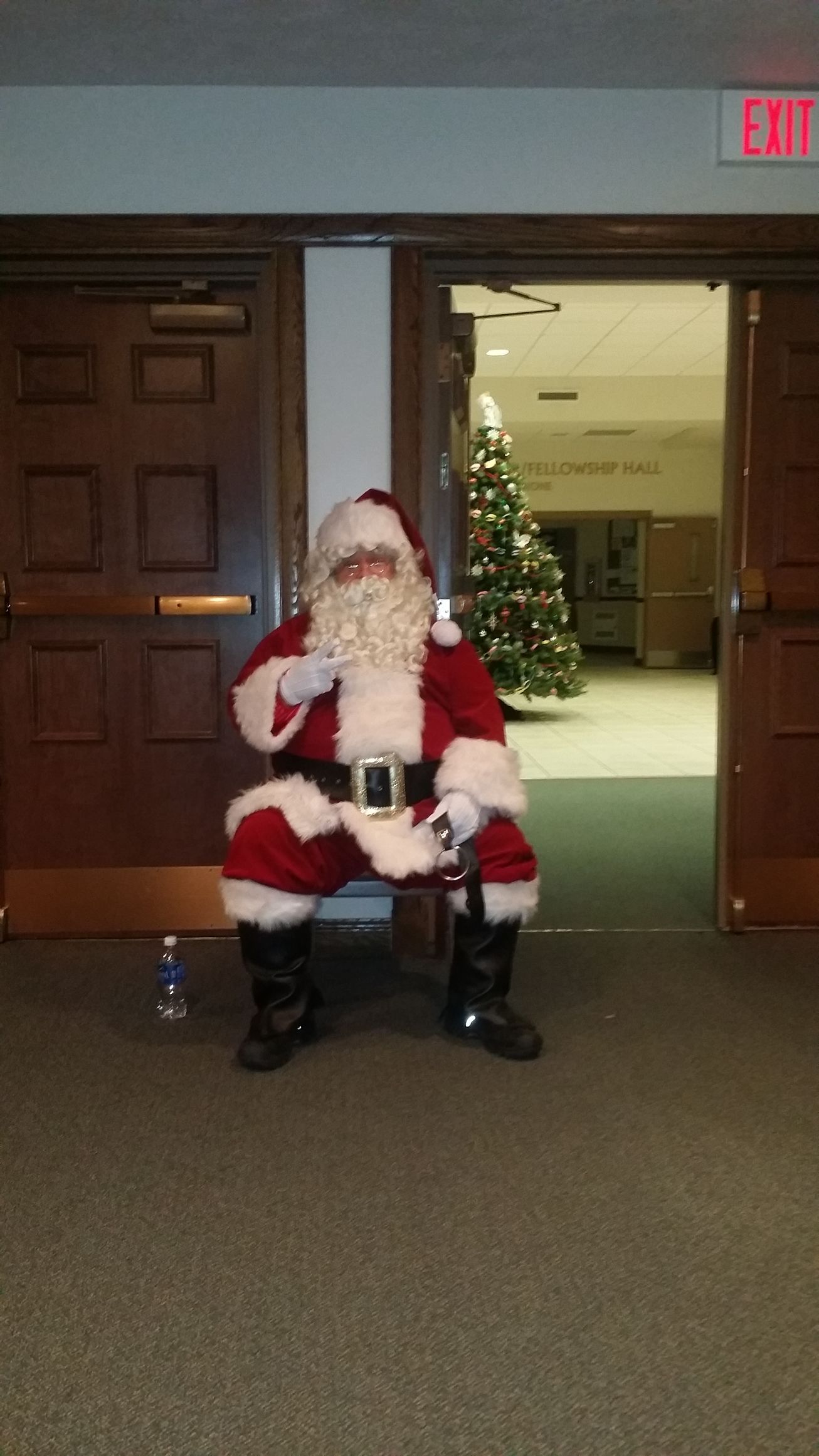 Santa Claus sitting on a chair in a hallway doorway, with a lit Christmas tree in the background.