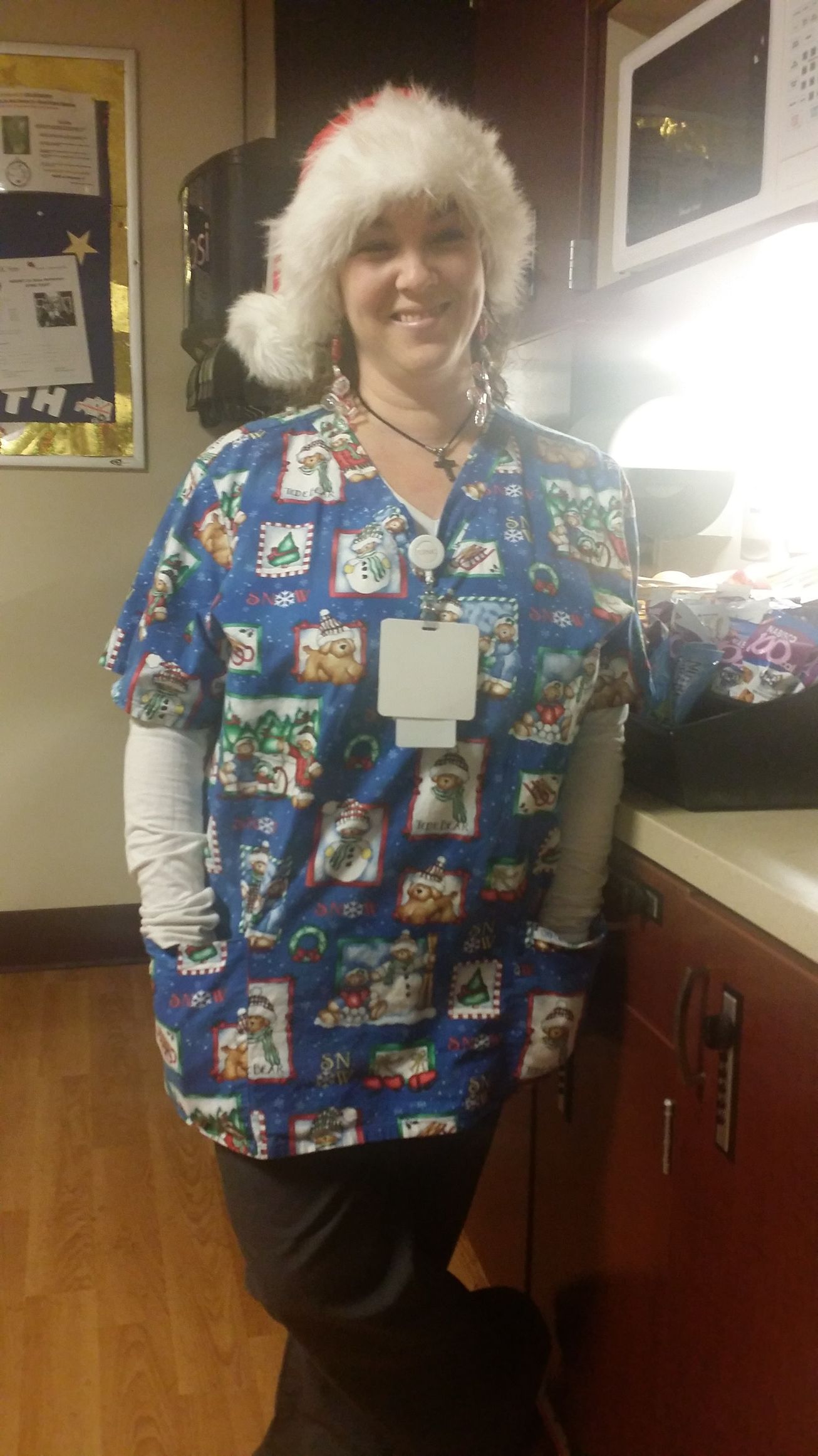 Person in blue patterned scrubs with white undershirt, standing in a kitchen and smiling.