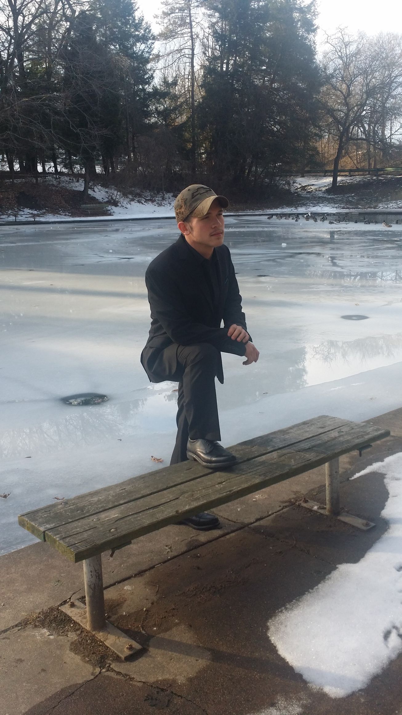 Person stands on a bench beside a frozen pond with snow on the ground and trees in the background.