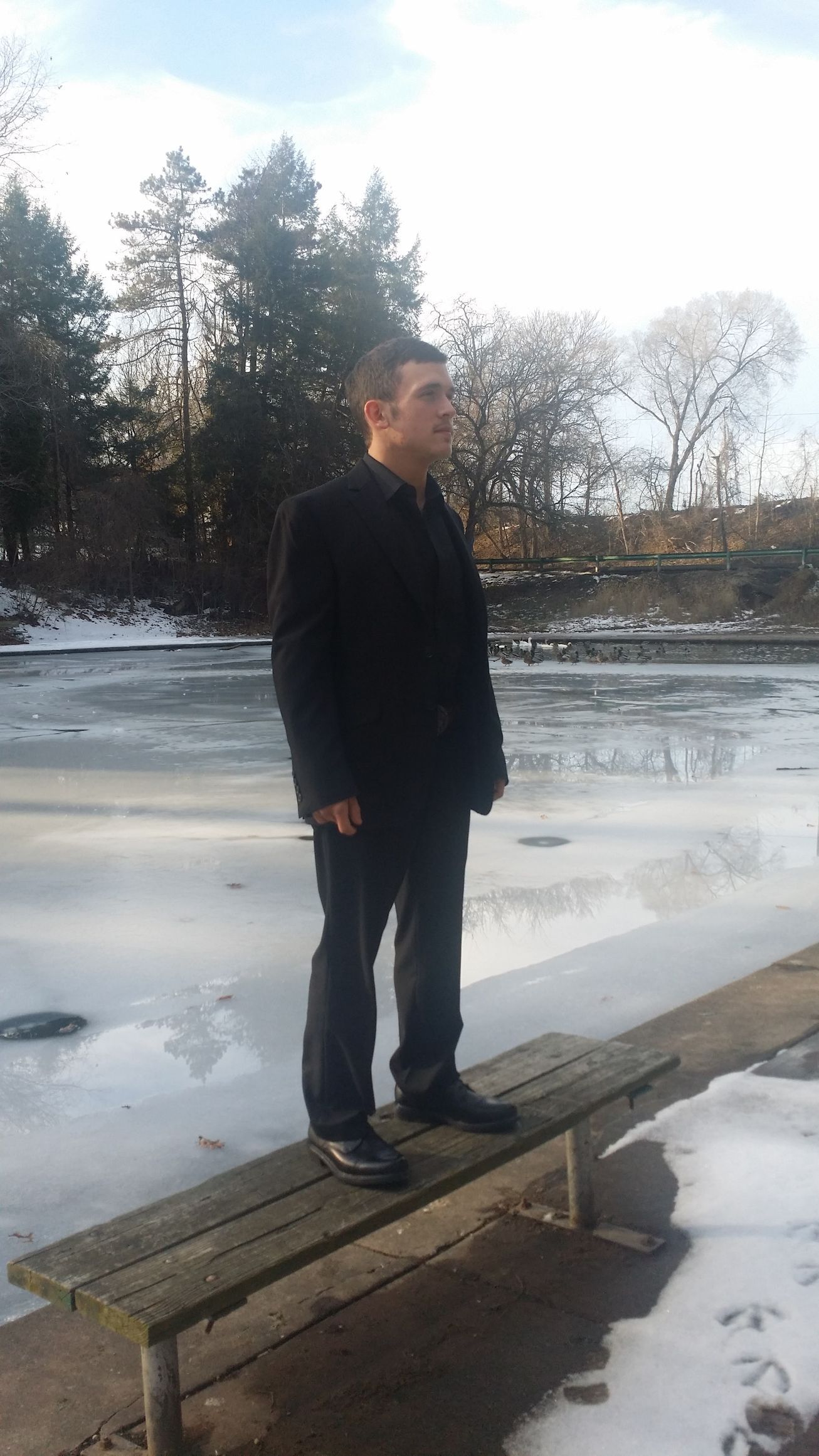 Man standing on a wooden bench by a frozen river, with trees and snow in the background