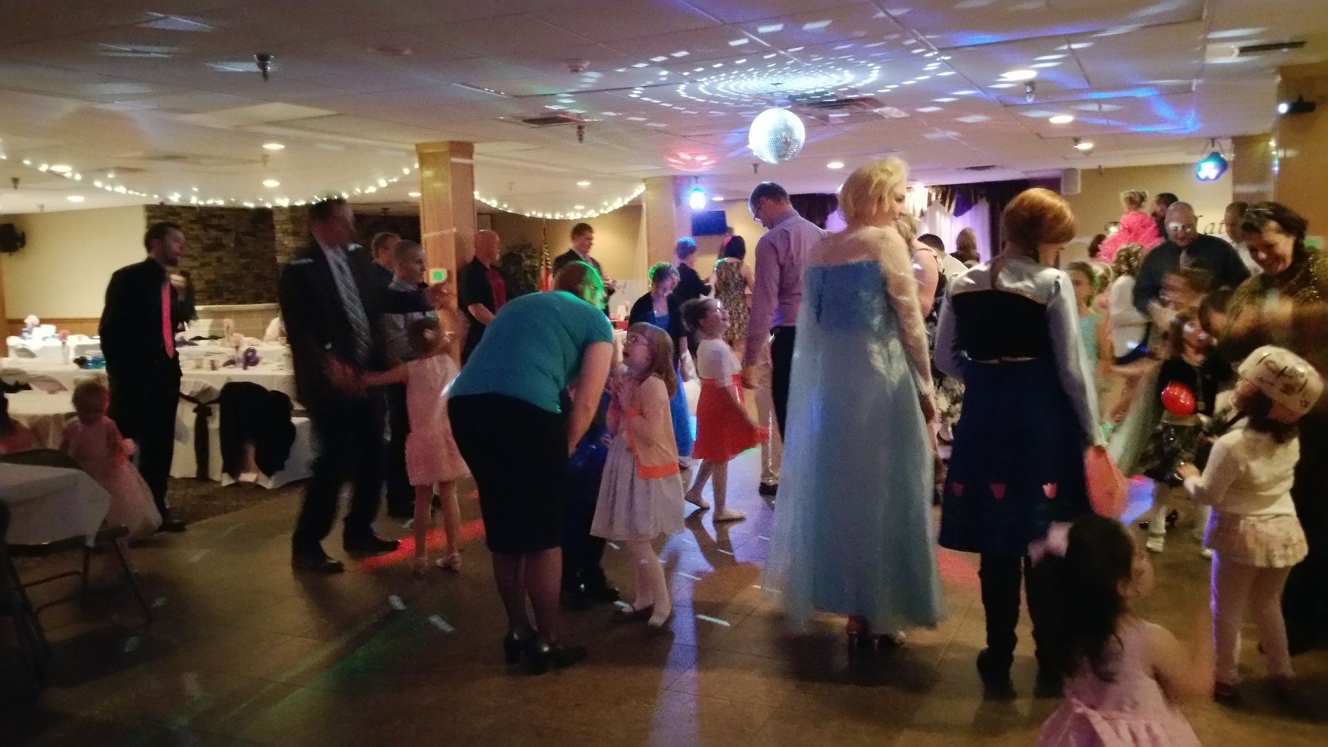 Guests dancing at a brightly lit indoor party, with colorful lights and a child in a white dress nearby