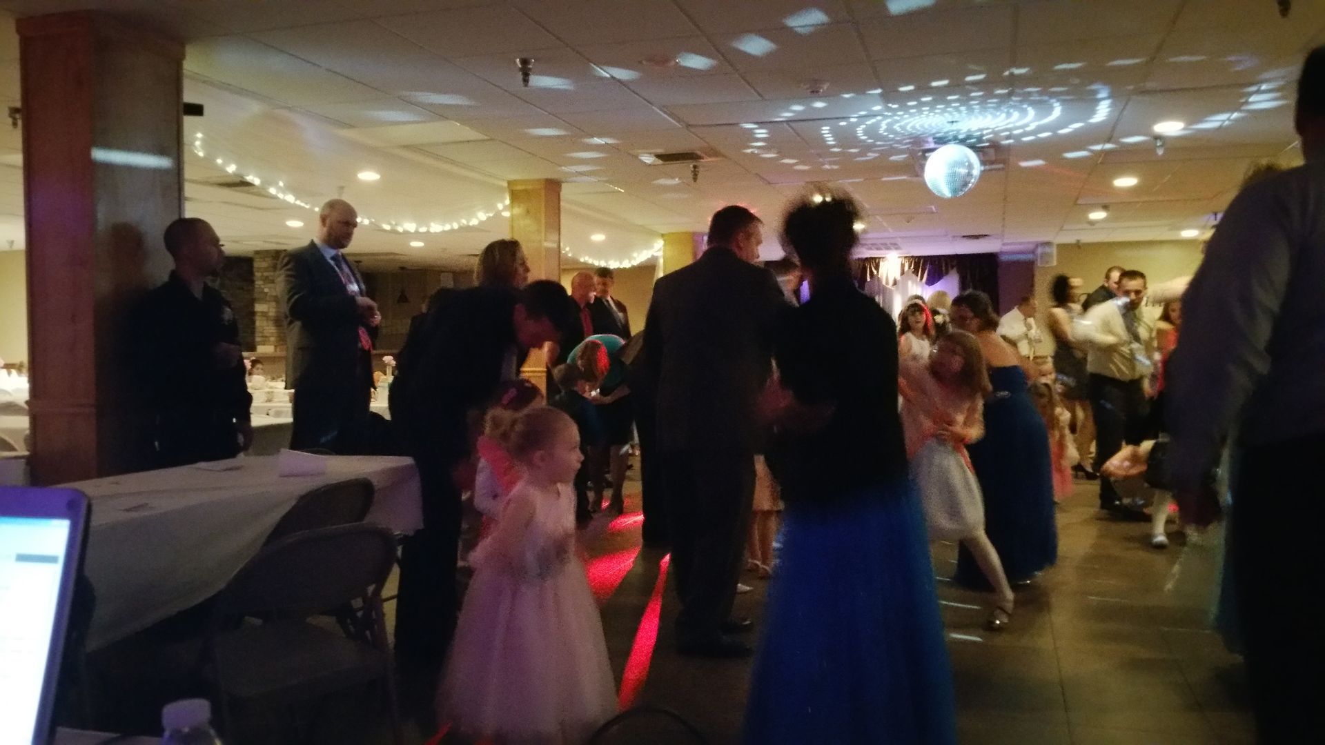 Guests dancing at a warmly lit banquet hall, with colorful lights and a child in a white dress in the foreground