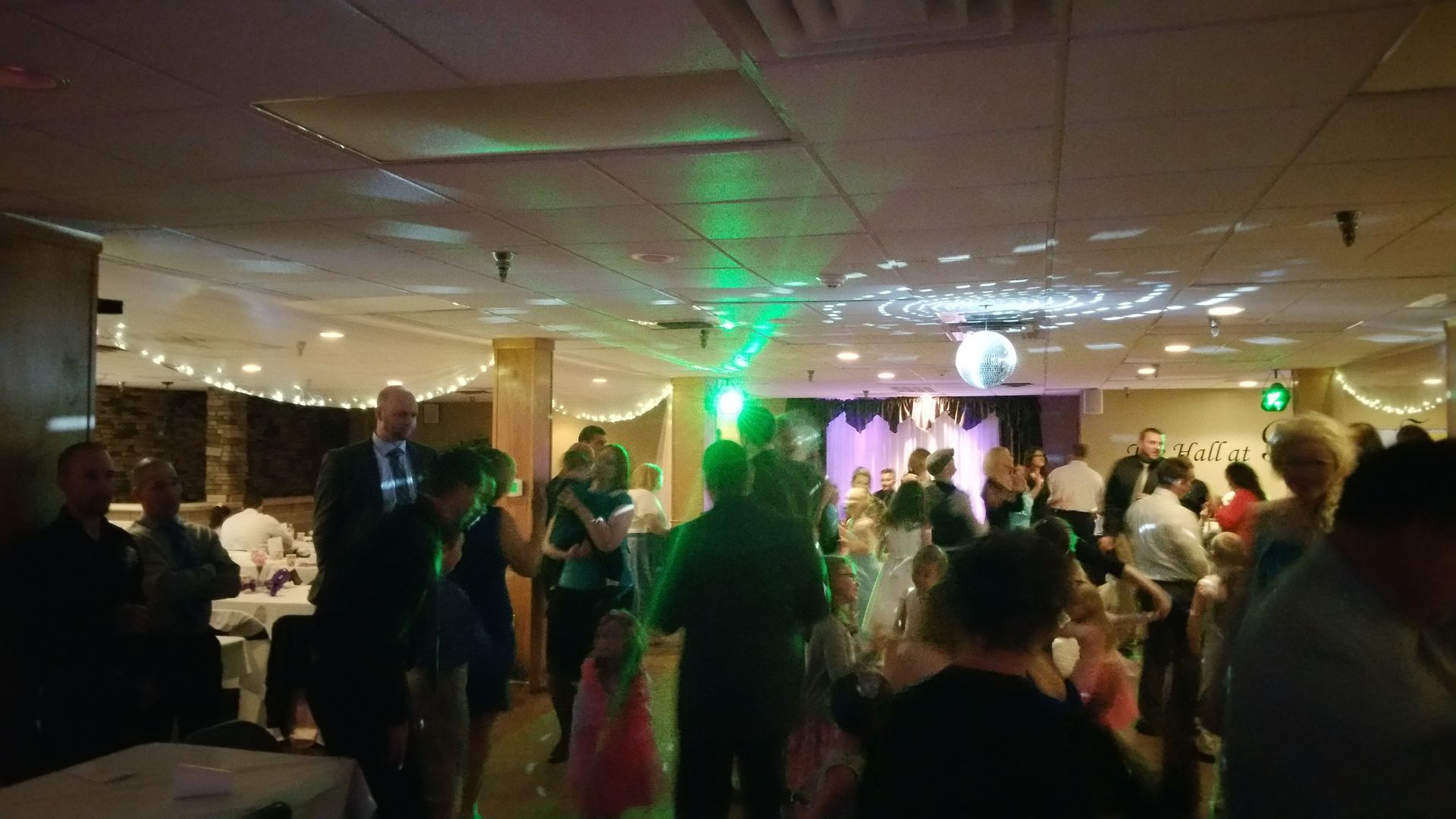 Crowded banquet hall with people dancing under green and purple lights near a stage