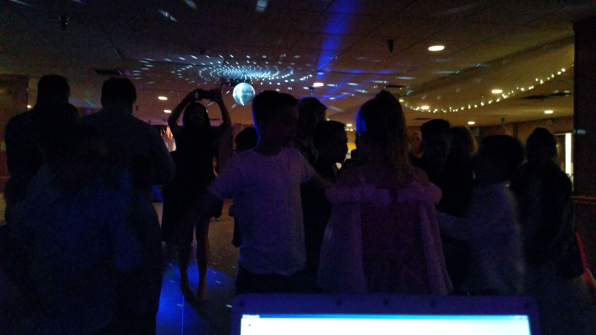Crowd standing in a dim room with blue stage lights and a glowing screen in front