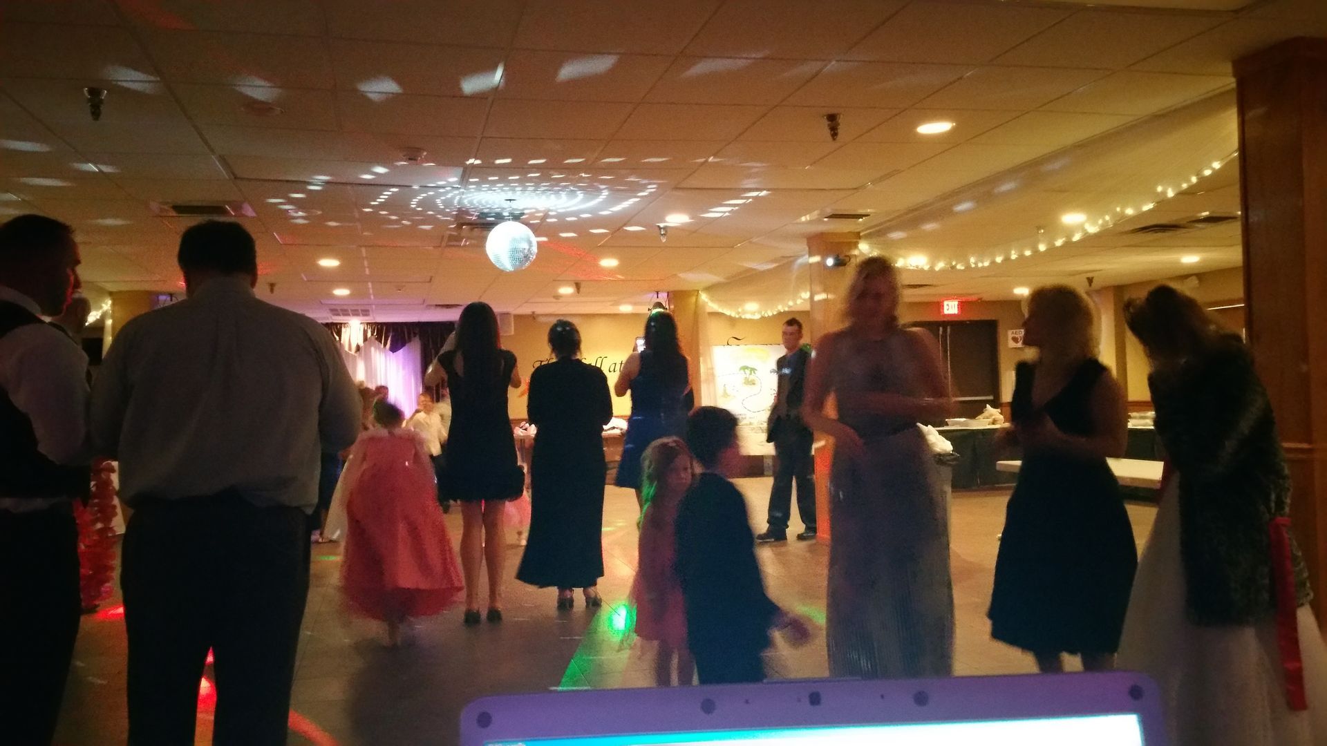 People dancing in a warmly lit banquet hall with colorful lights and a disco ball.