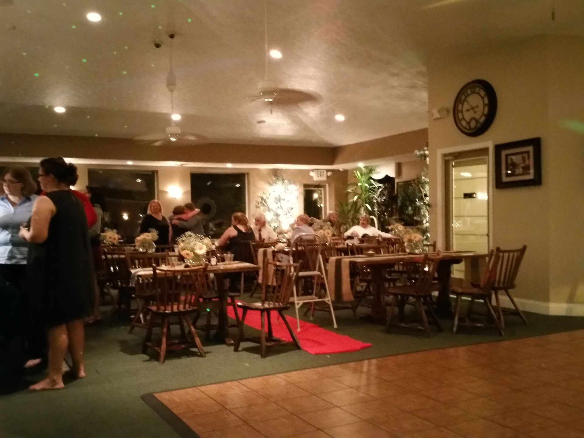 Restaurant dining room with diners seated at wooden tables under warm ceiling lights and a red carpeted aisle