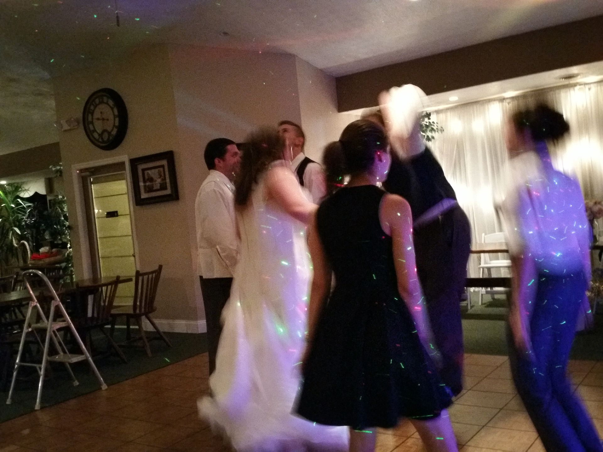 People dancing at a dimly lit indoor party, with colorful lights and chairs along the wall.