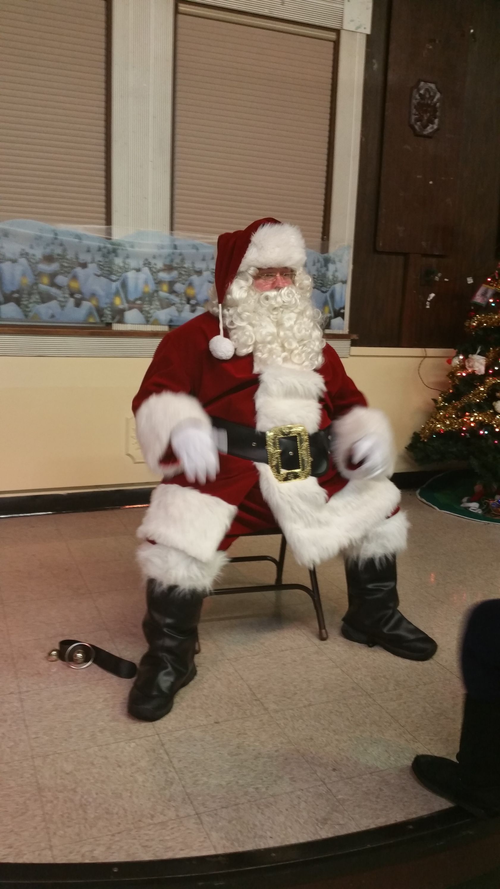 Santa Claus seated indoors, wearing a red suit and black boots, with a Christmas tree in the background