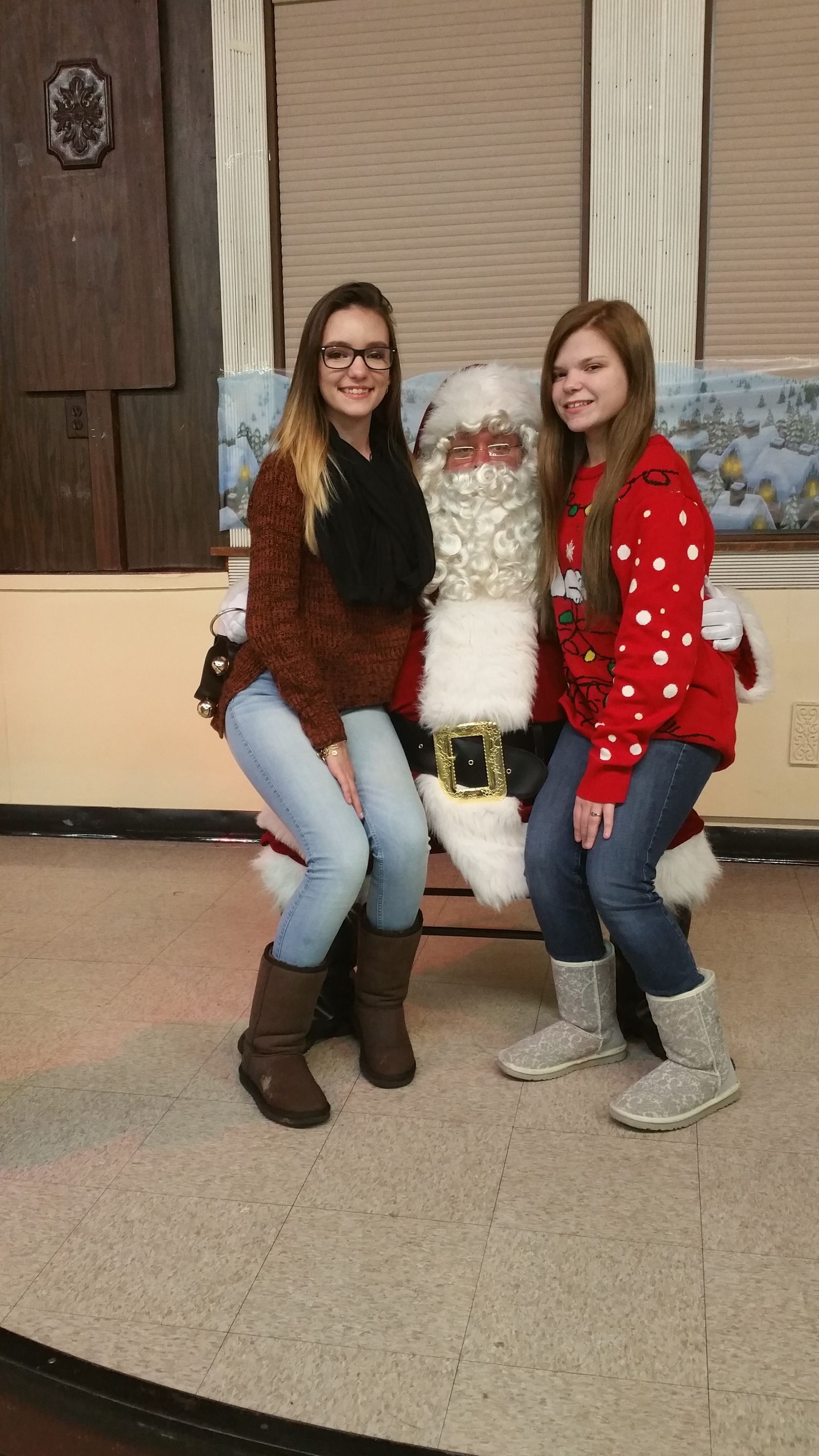 Two women kneel beside a Christmas tree display in a decorated indoor room, smiling at the camera.