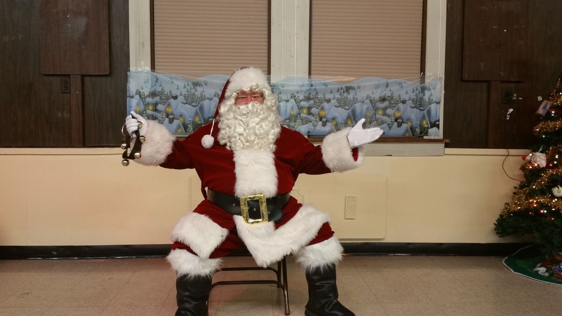 Santa Claus seated indoors with arms open, wearing a red suit and white beard, against a holiday backdrop