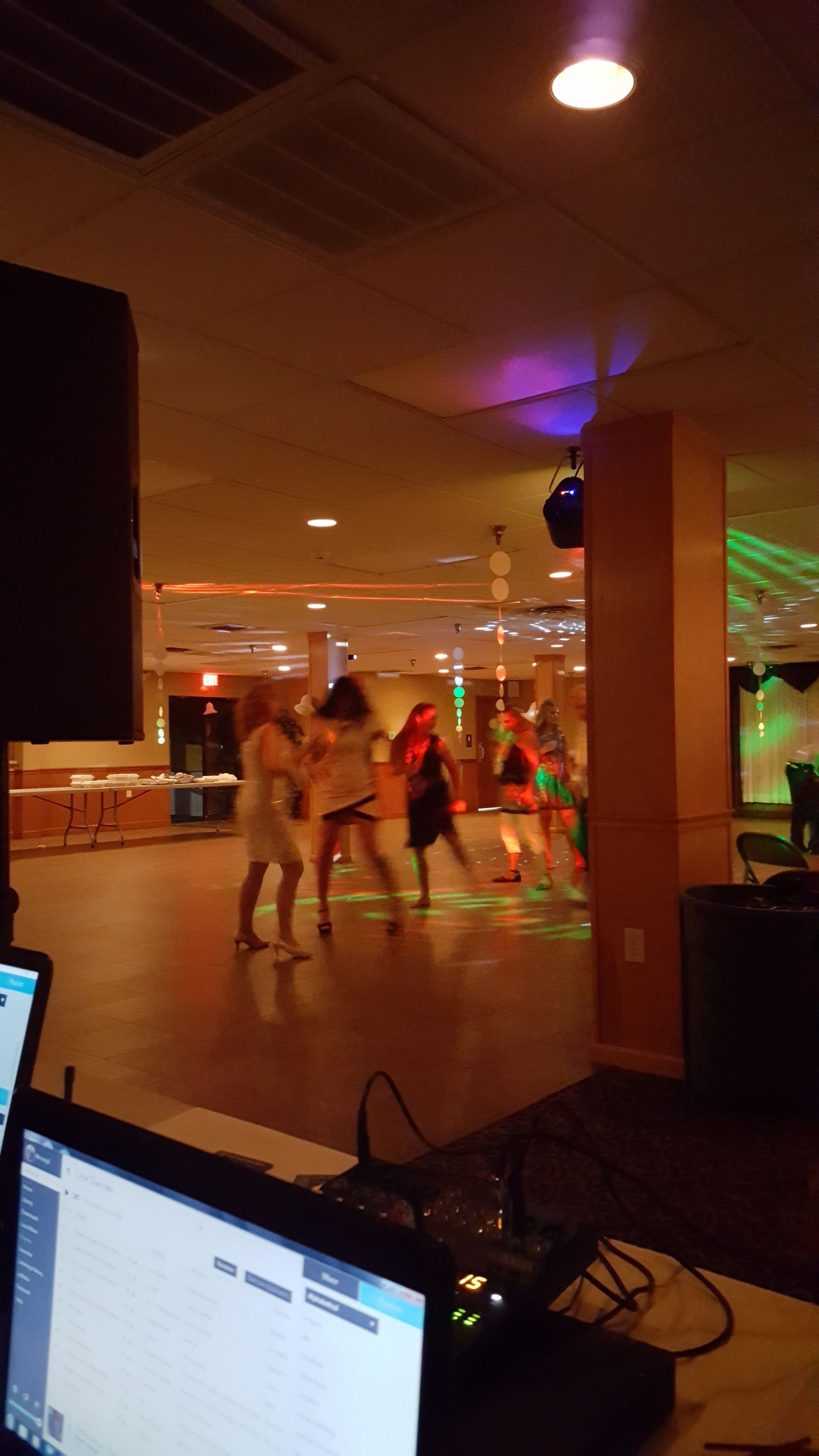 Dimly lit indoor dance floor with people dancing, colorful lights, and a laptop on a table in the foreground