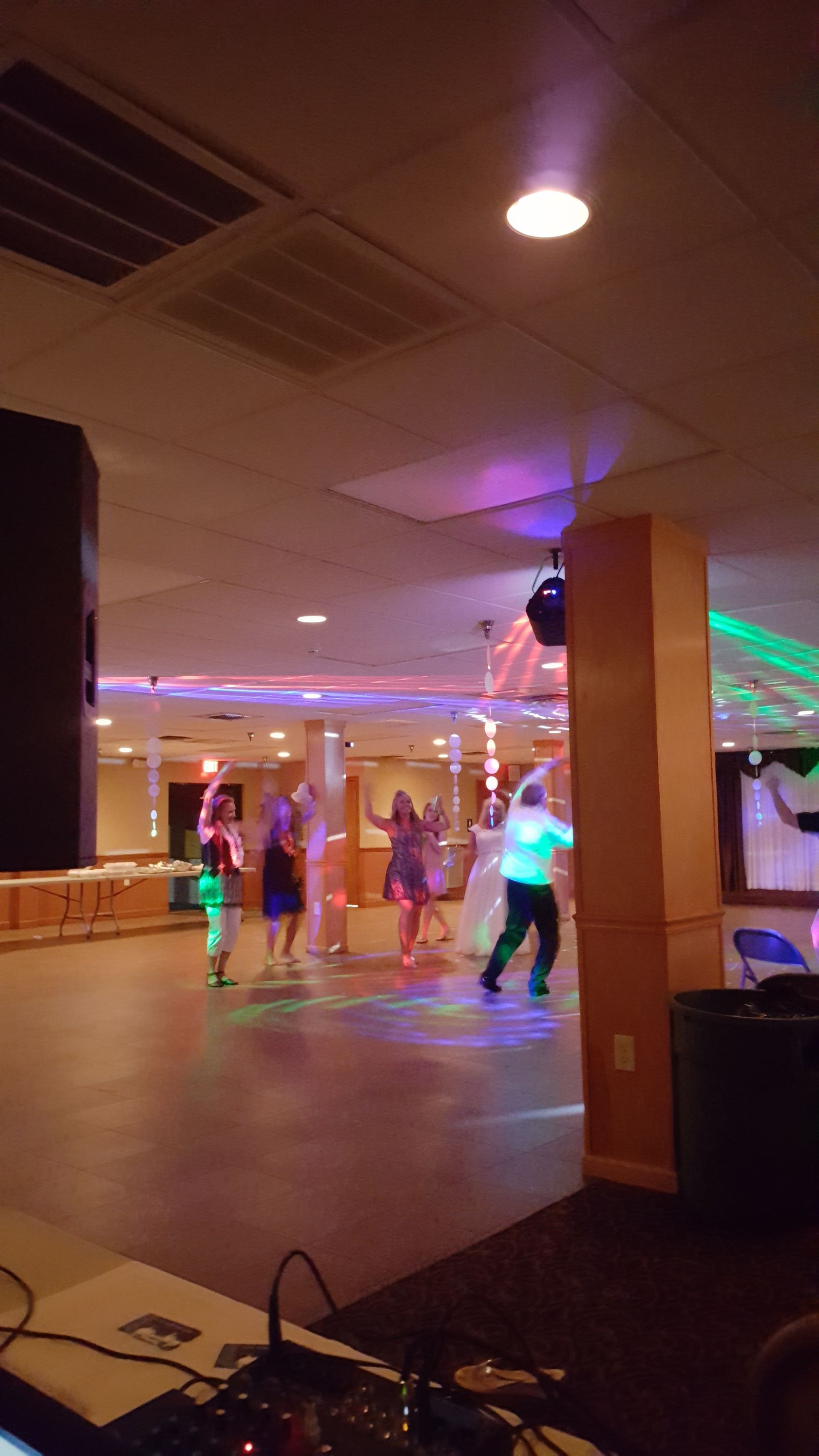 People dancing in a dimly lit hall with colorful party lights and a speaker in the foreground