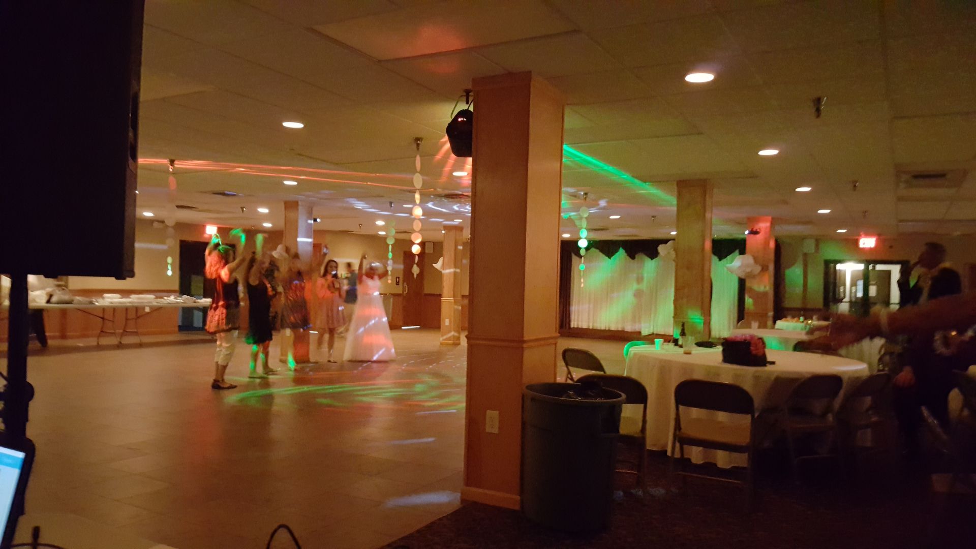 People dancing in a dimly lit banquet hall with green and orange lights, tables along the side.