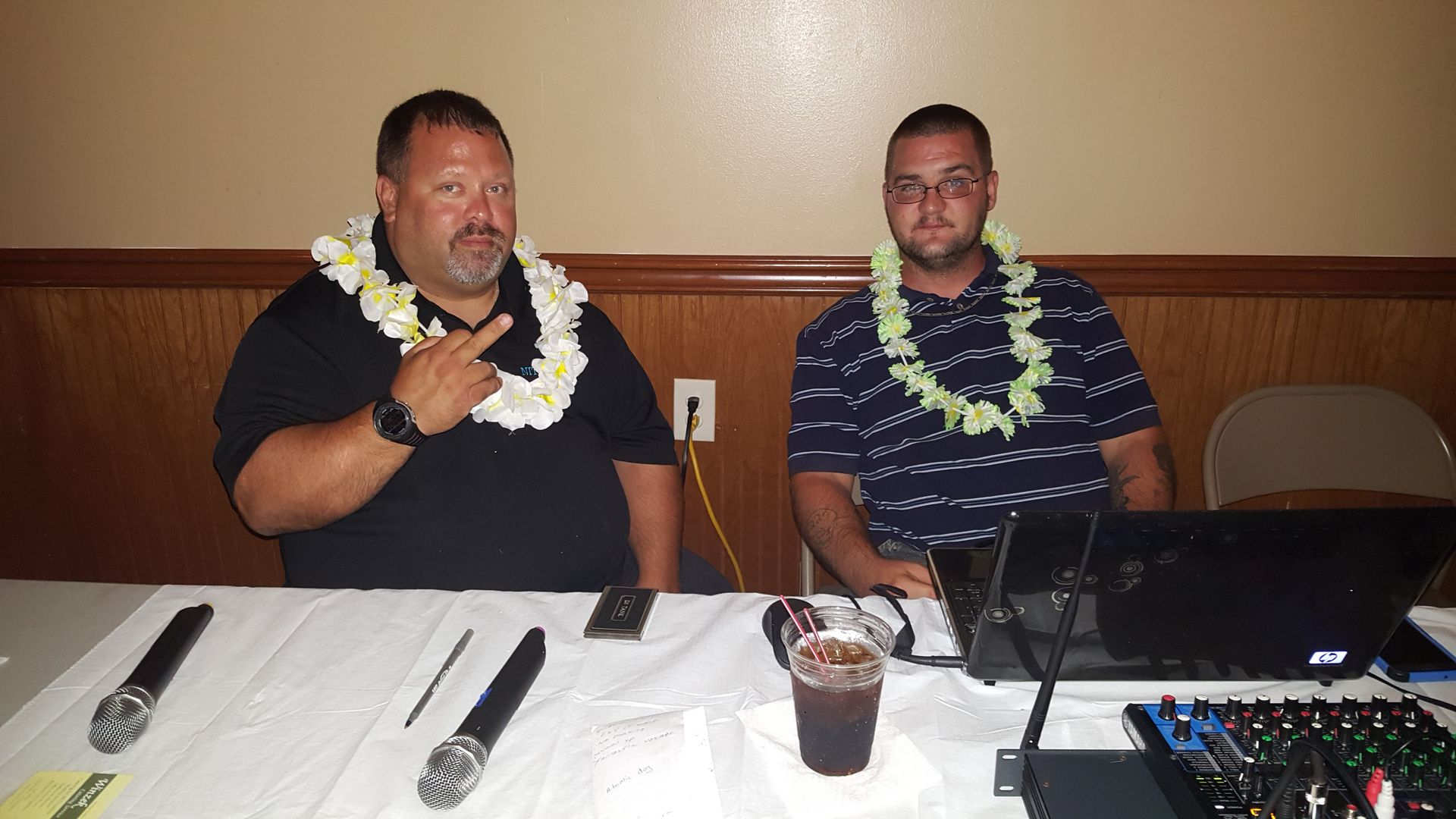 Two men seated at a banquet table with leis, one speaking into a microphone beside a laptop and audio equipment.