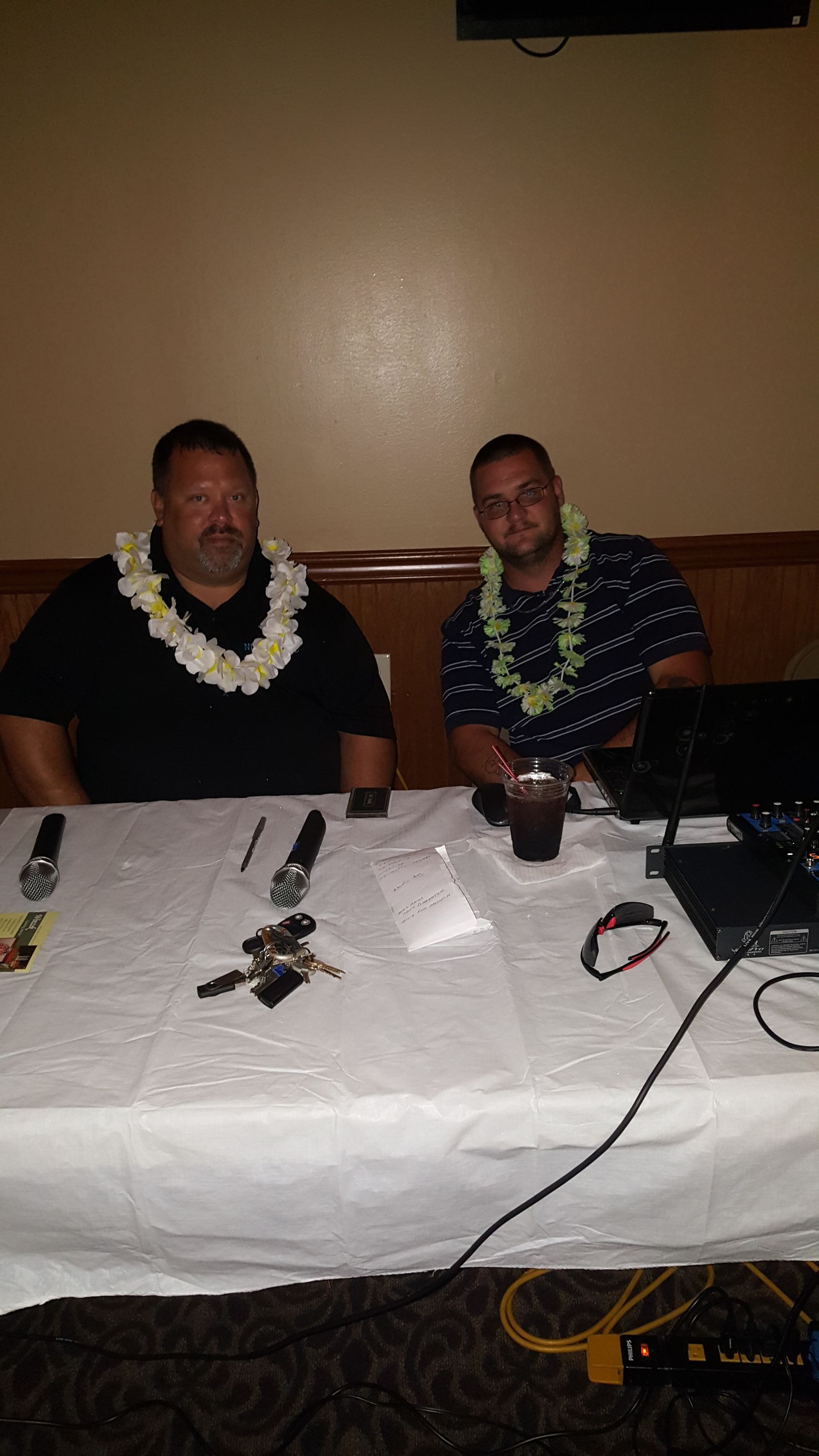Two people seated behind a white tablecloth at a dimly lit event, wearing lei garlands, with audio equipment on the table