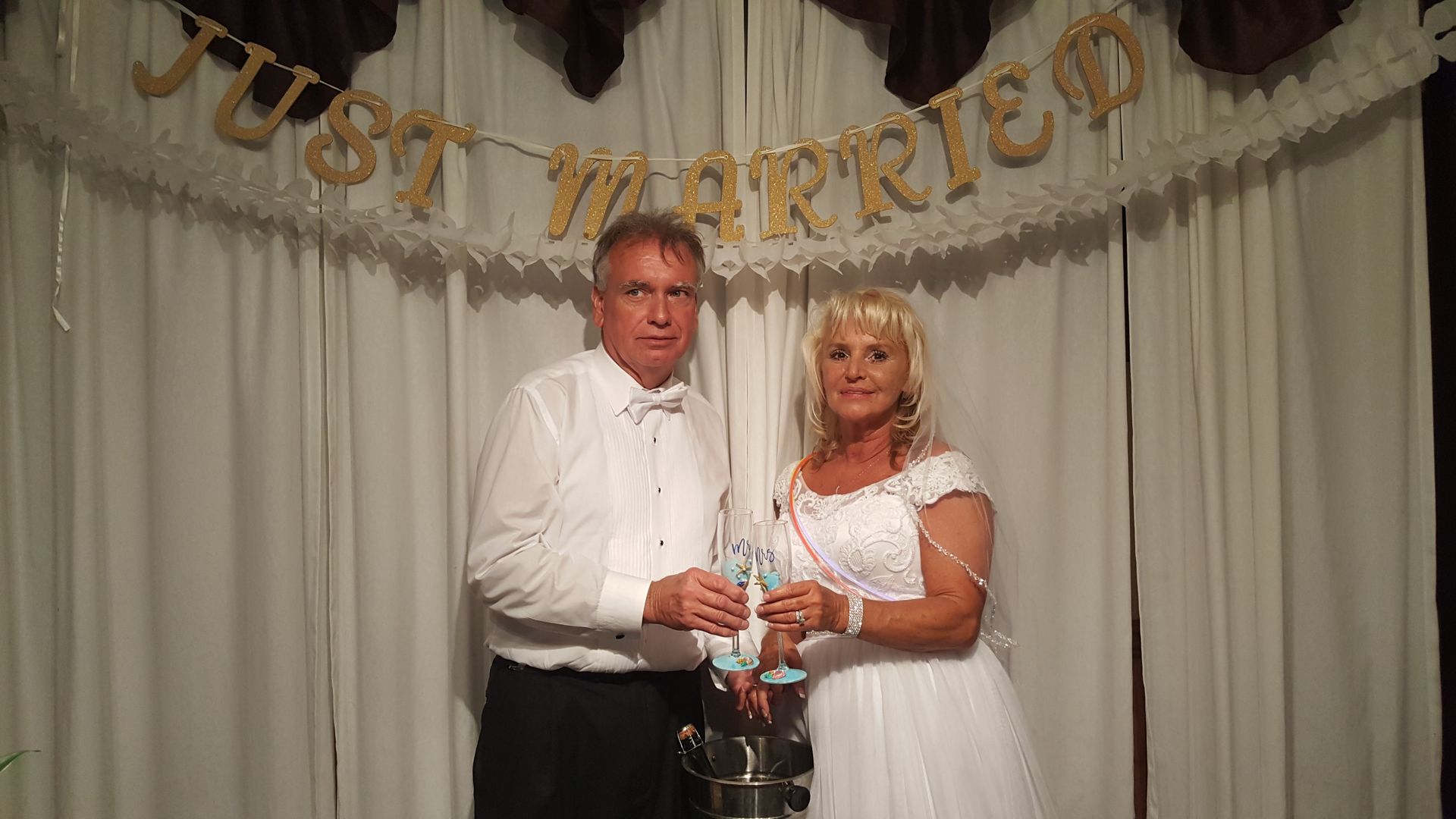 Couple in white outfits holding a silver goblet at an anniversary celebration backdrop