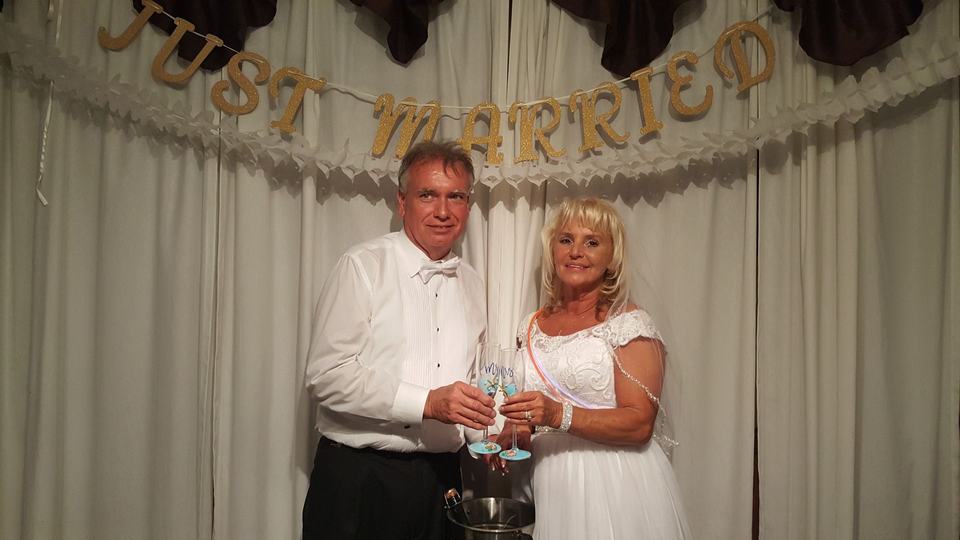 Two people in white outfits holding a trophy in front of a “Just Married” banner