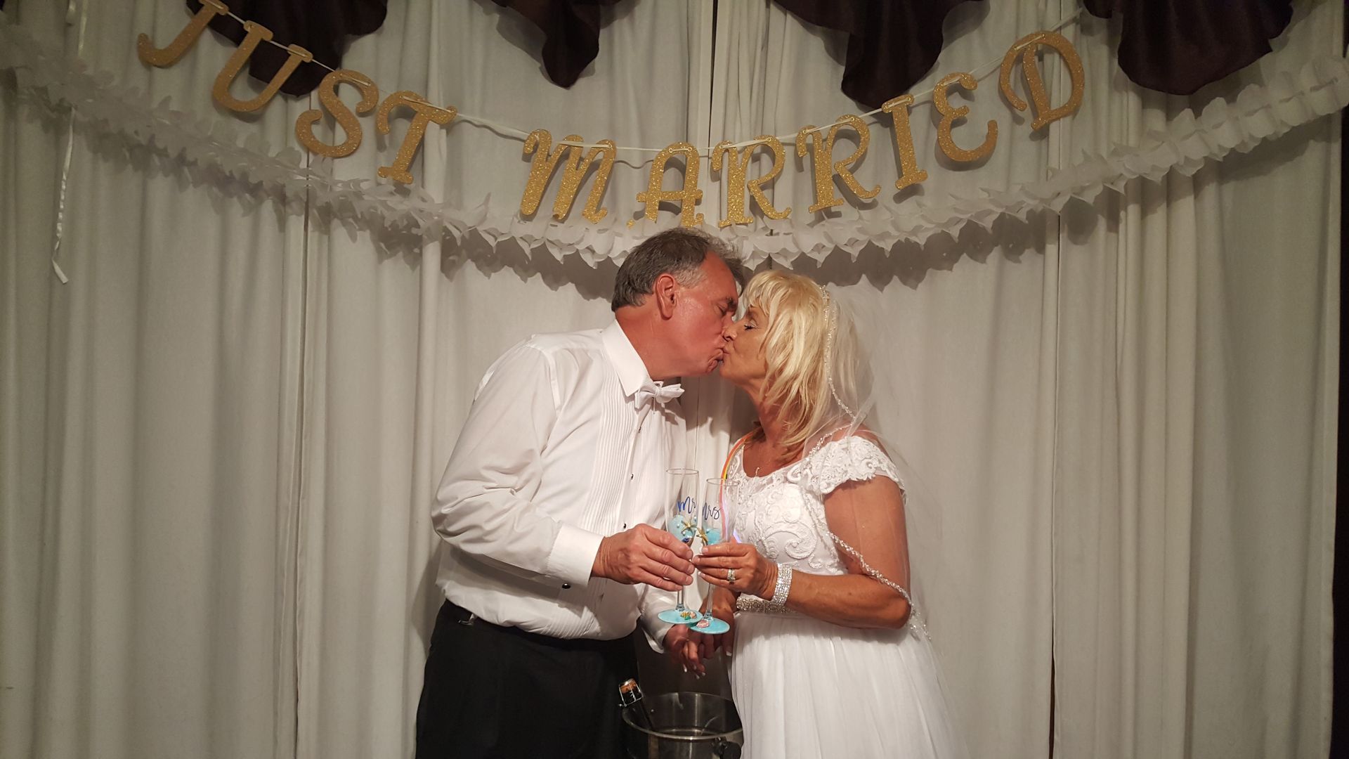 Newlyweds kissing while holding champagne glasses under a “Just Married” banner