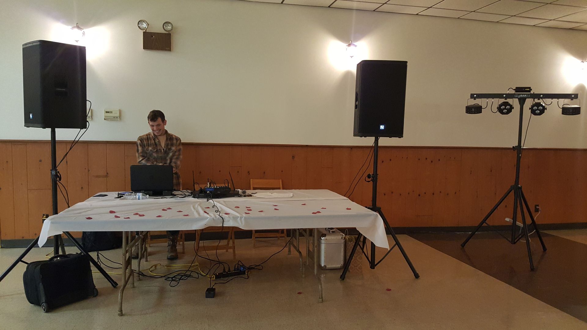 DJ setup in a hall with two speakers, a mixer table, and a person behind equipment on a small stage