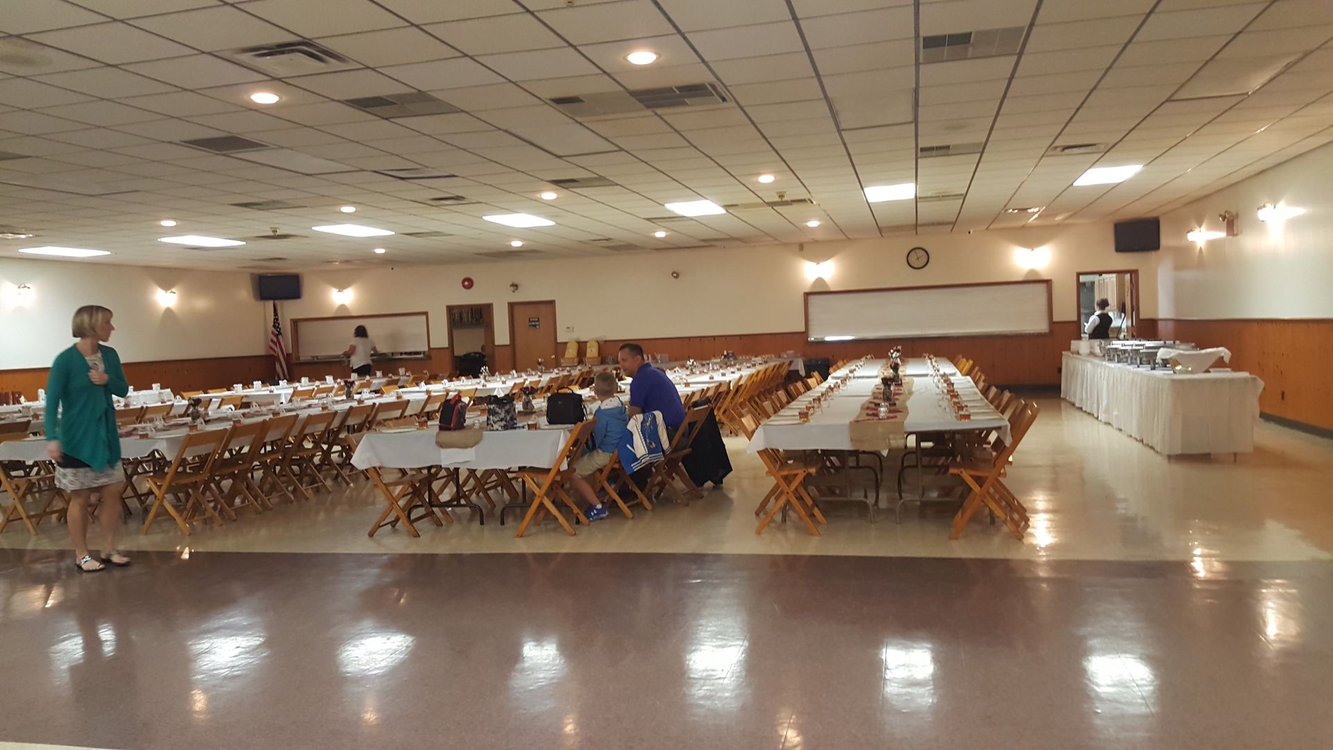 Large banquet hall with rows of tables and chairs, a few people setting up, and a buffet table along the wall.