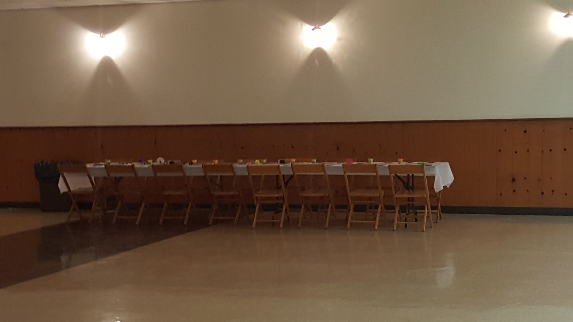 Empty banquet hall with a long table and rows of folding chairs against a wood-paneled wall.