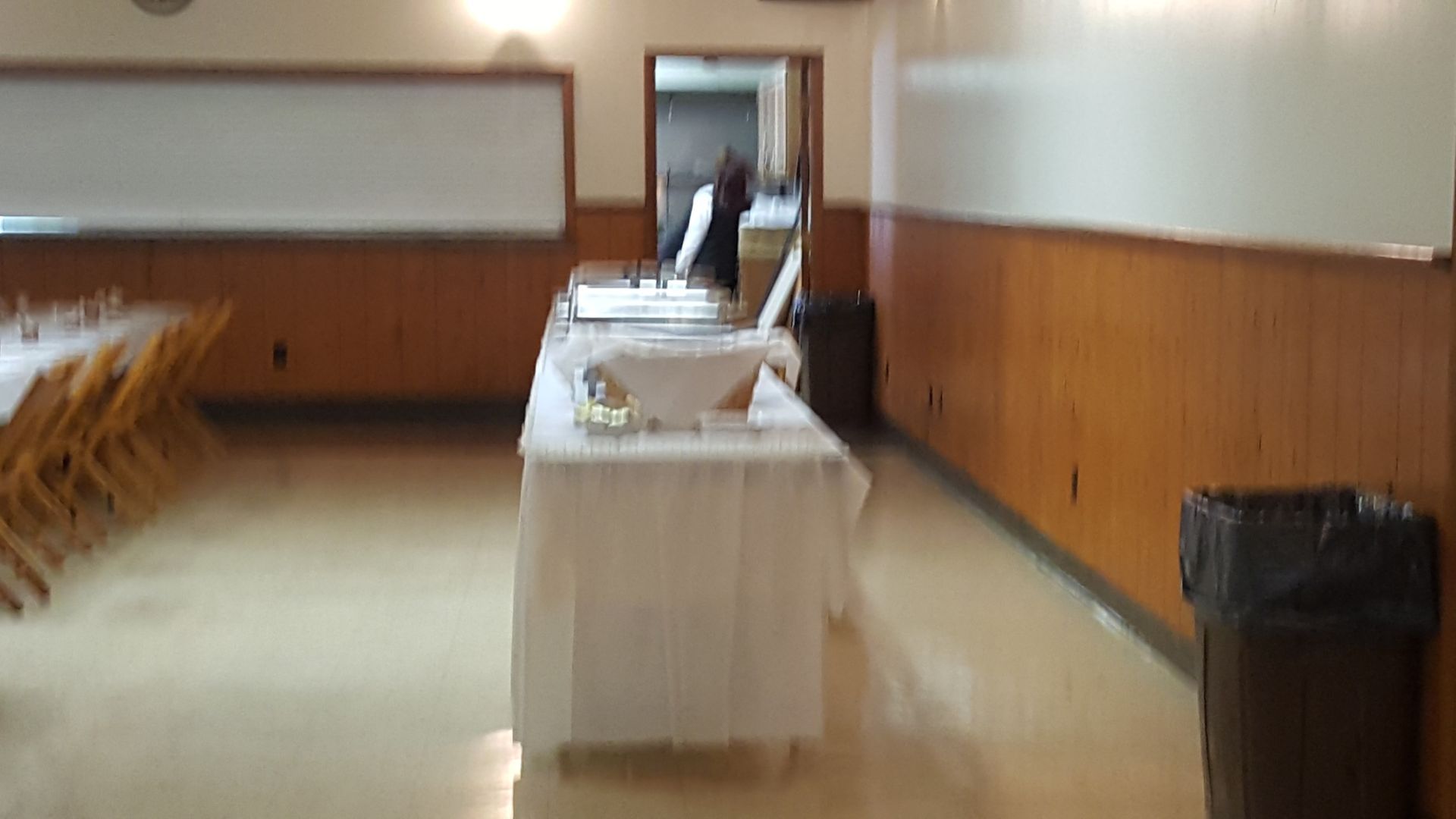 Buffet table in a wood-paneled hall with chairs along the left wall and a trash can on the right.