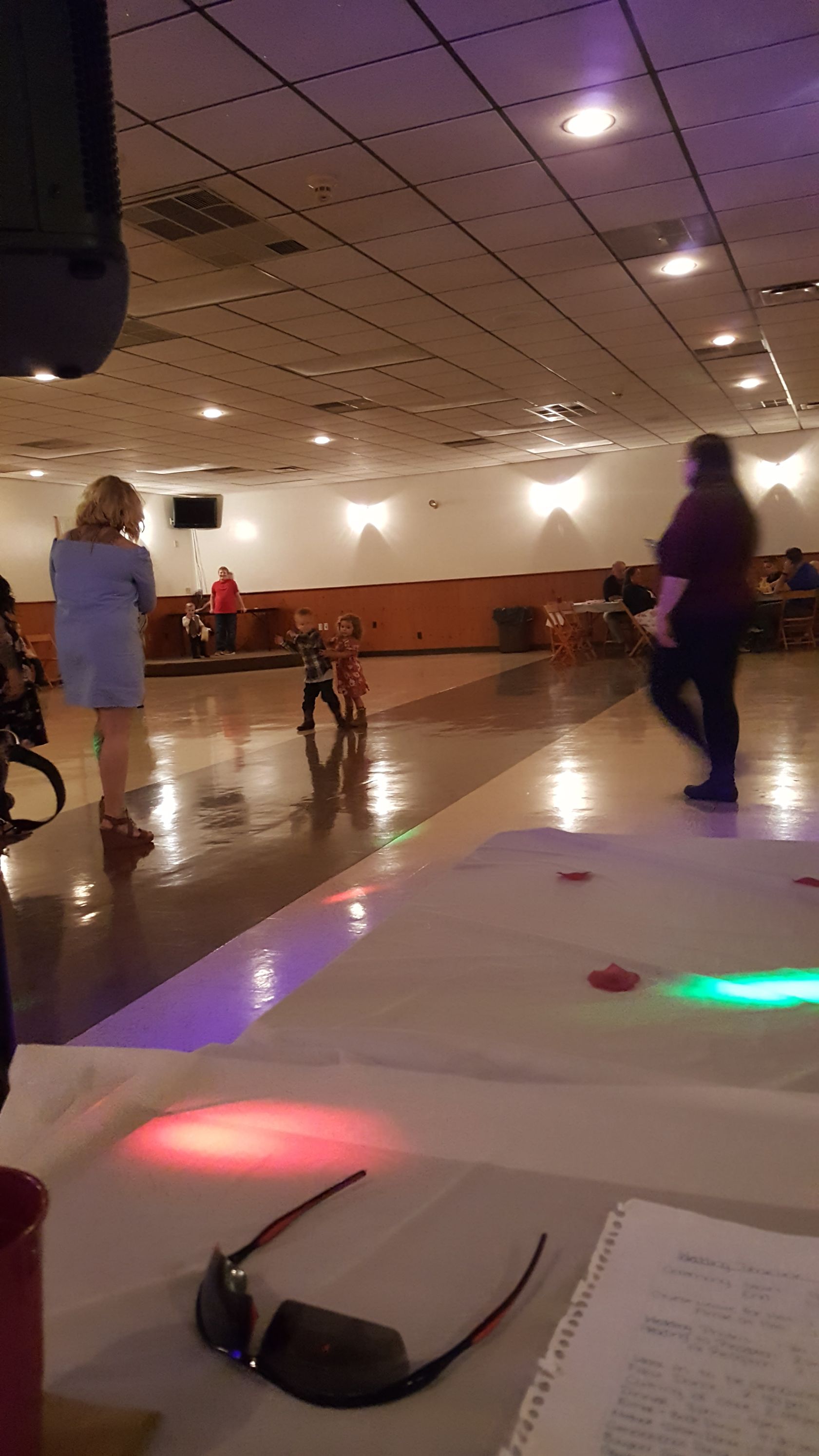 People dancing in a banquet hall with reflective floor and ceiling lights, viewed from a table with sunglasses.