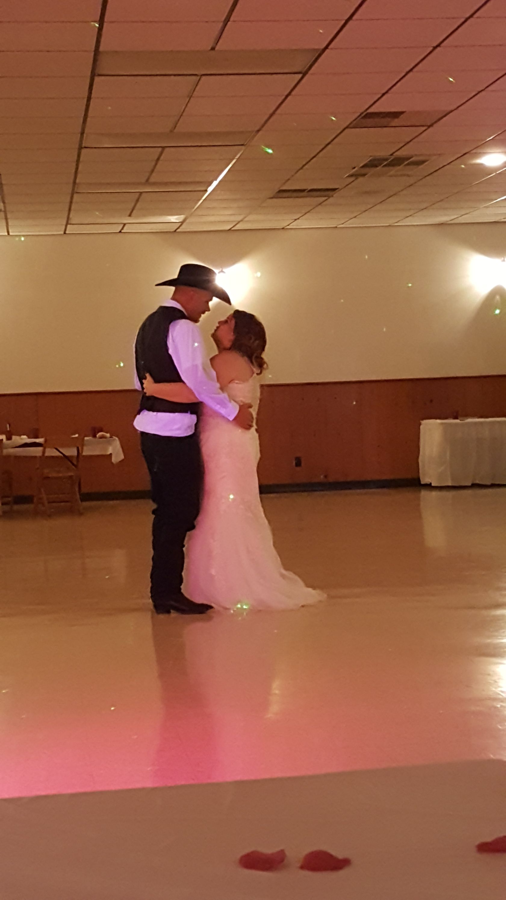 Couple slow dancing in a banquet hall, the woman in a light pink dress and the man in a cowboy hat.