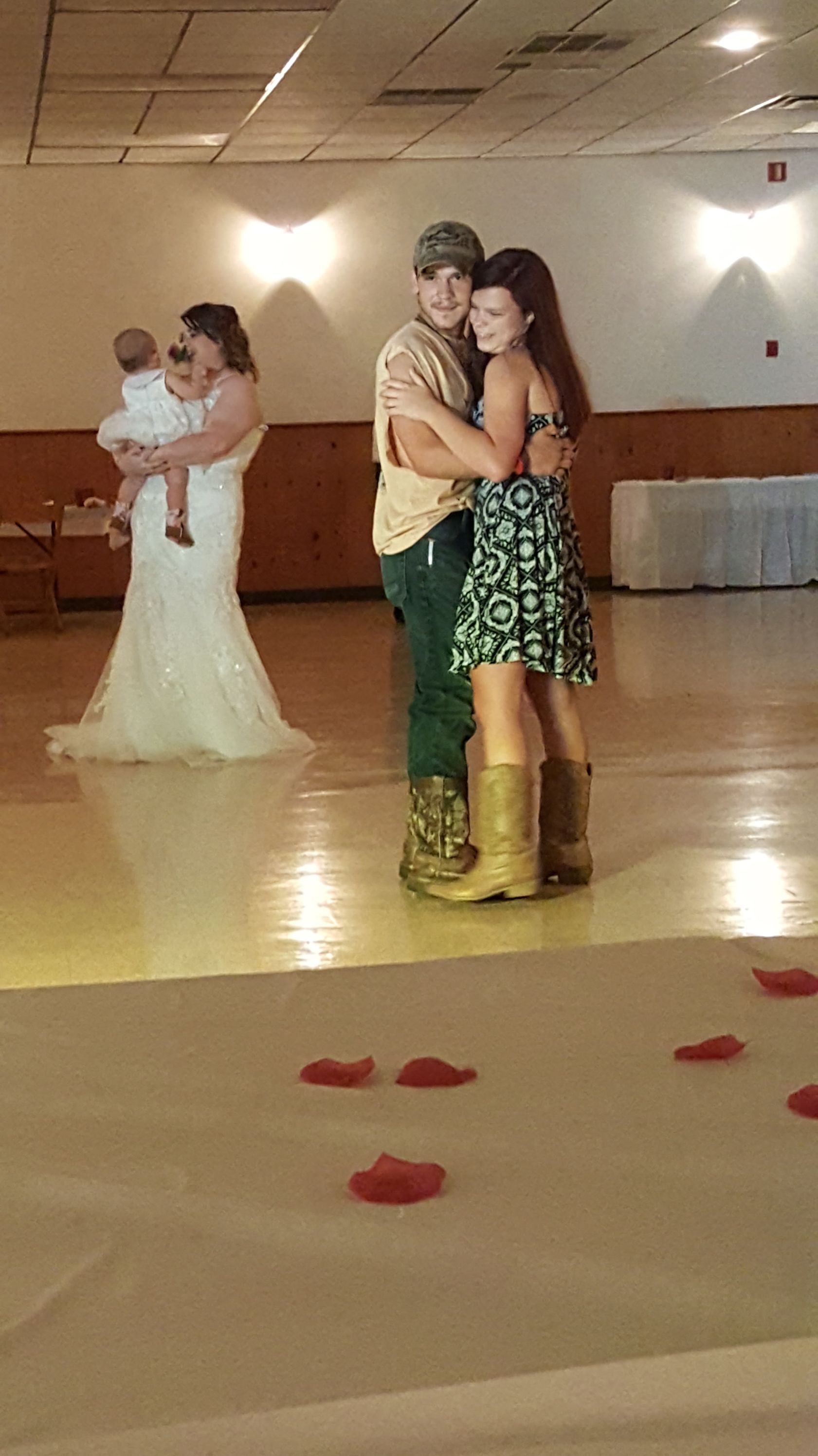Two children hugging on a dance floor with rose petals in the foreground; another couple dances in the background.