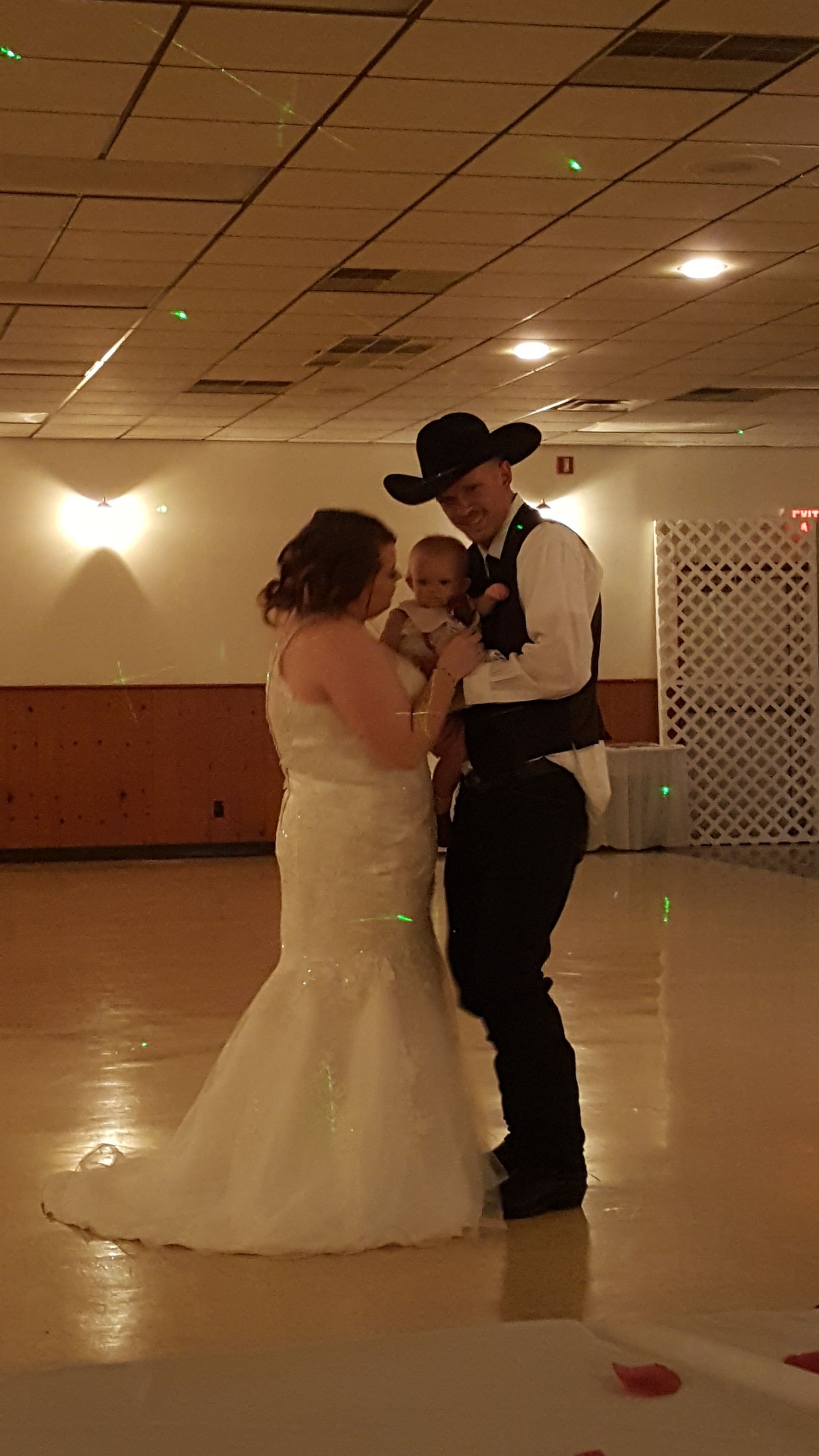 Couple slow dancing at a reception hall, woman in white gown and man in black hat and vest.