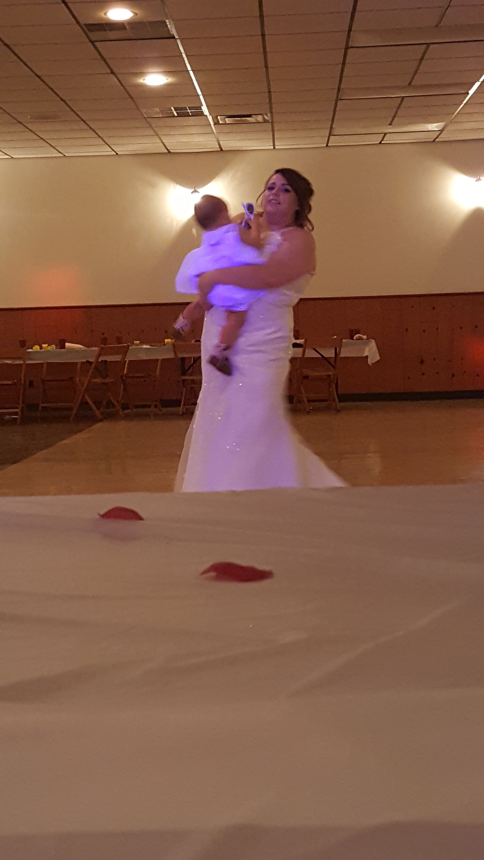 Bride in a white dress holding a baby under purple light at a reception hall