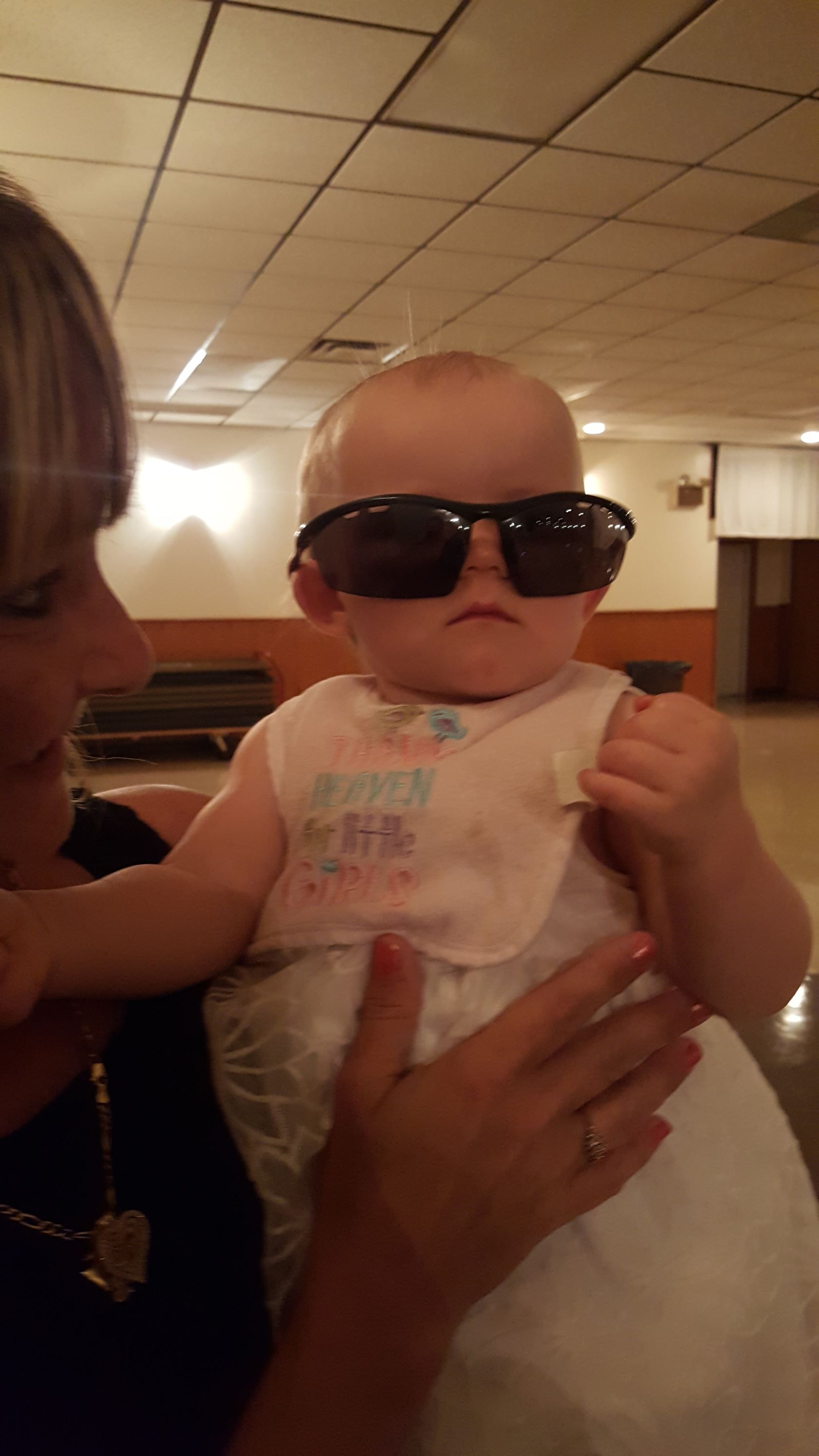 Baby wearing oversized sunglasses being held indoors under warm ceiling lights