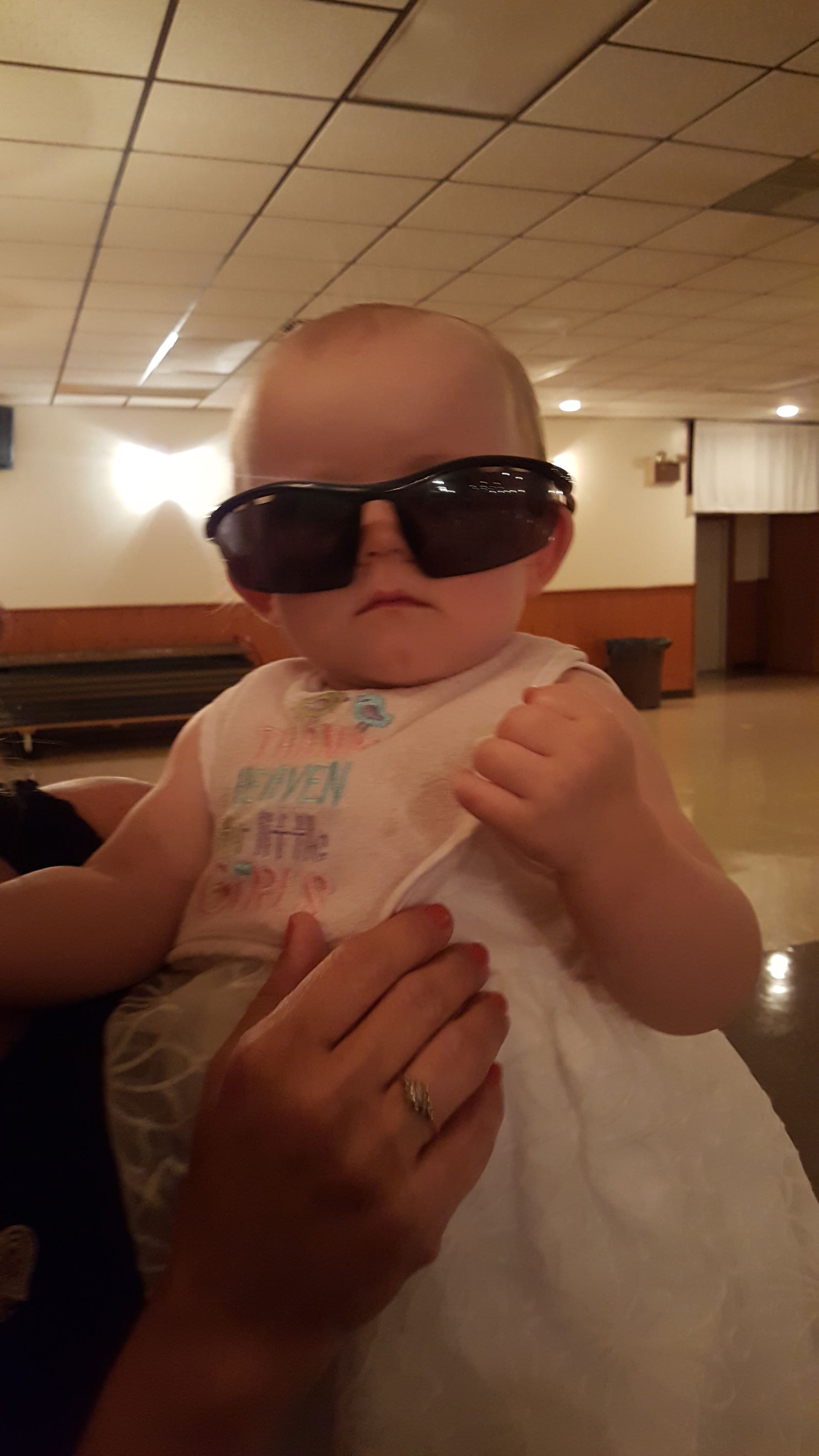 Baby in oversized sunglasses being held indoors under warm ceiling lights.