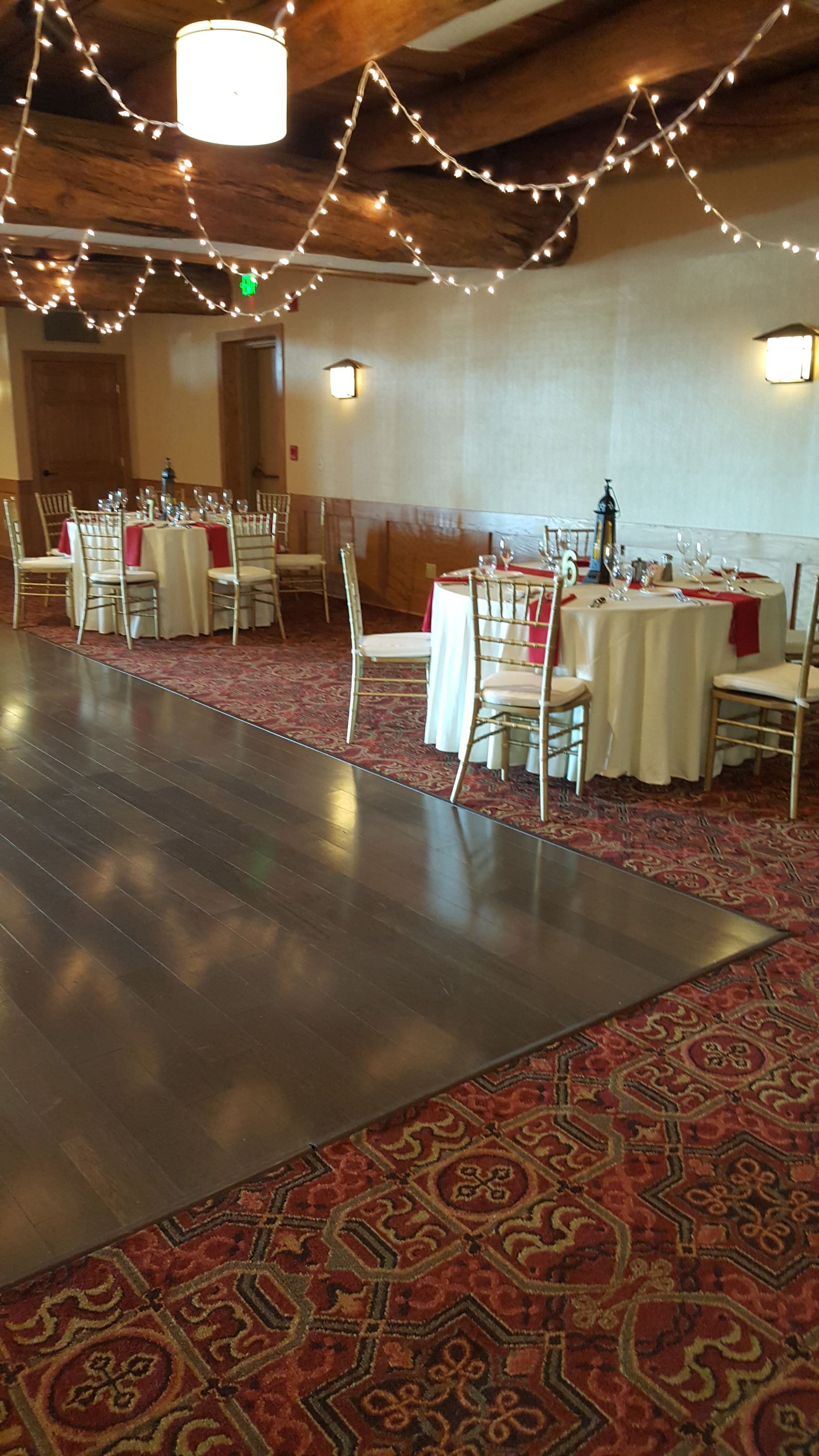 Decorated banquet hall with round tables, white chairs, string lights, and red patterned carpet