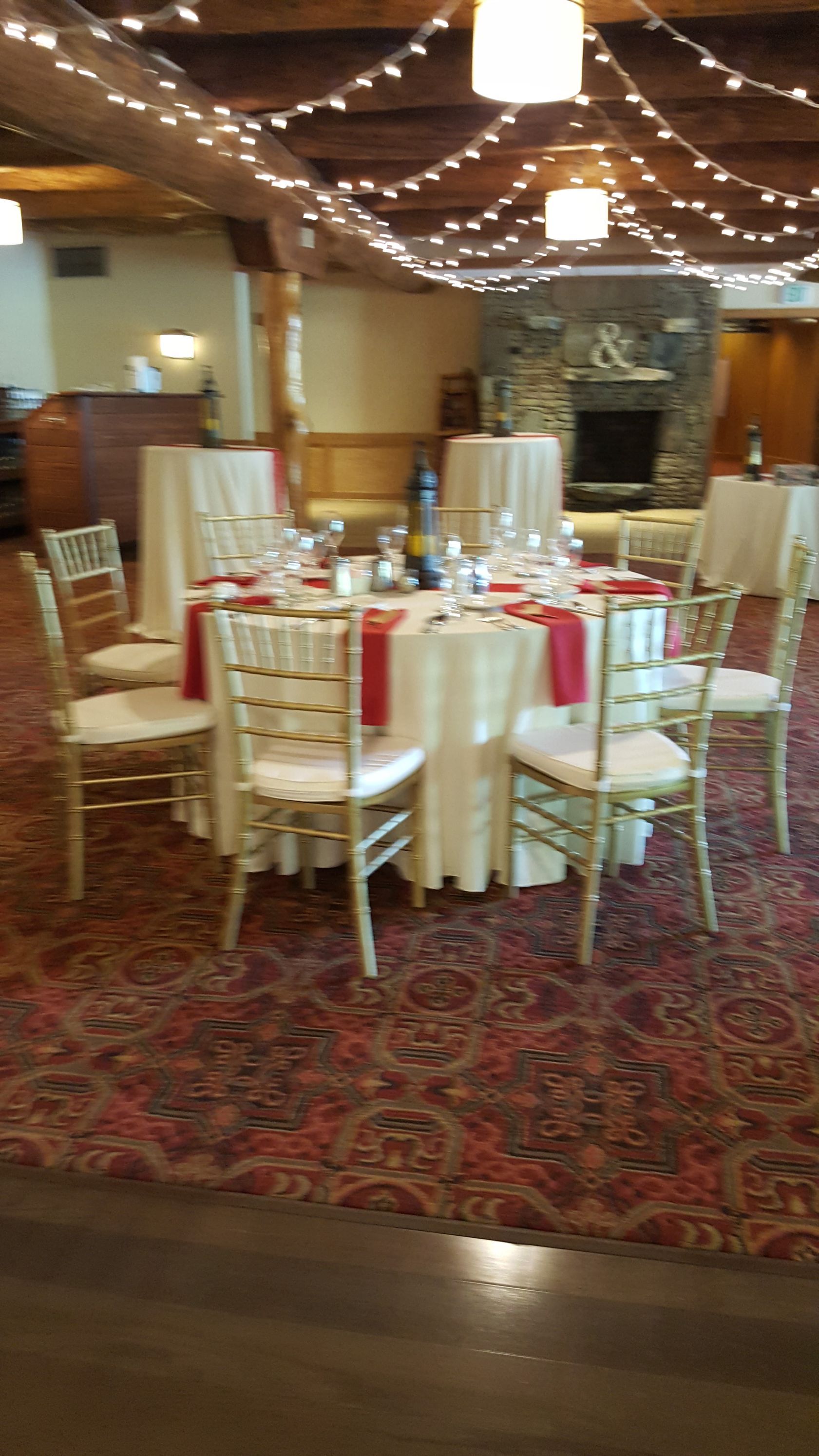 Decorated banquet hall with round tables, white chairs, red accents, and string lights overhead