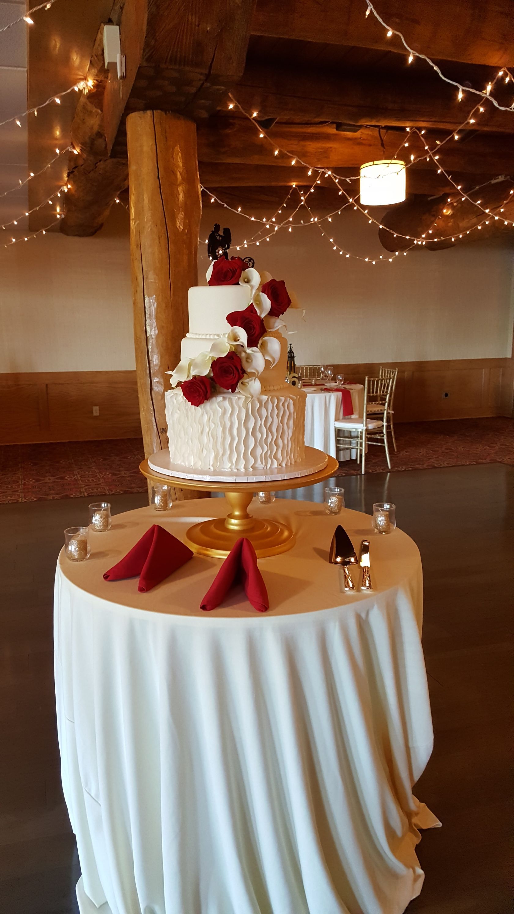 Wedding cake on a white-draped table with red and white flowers in a warmly lit rustic venue