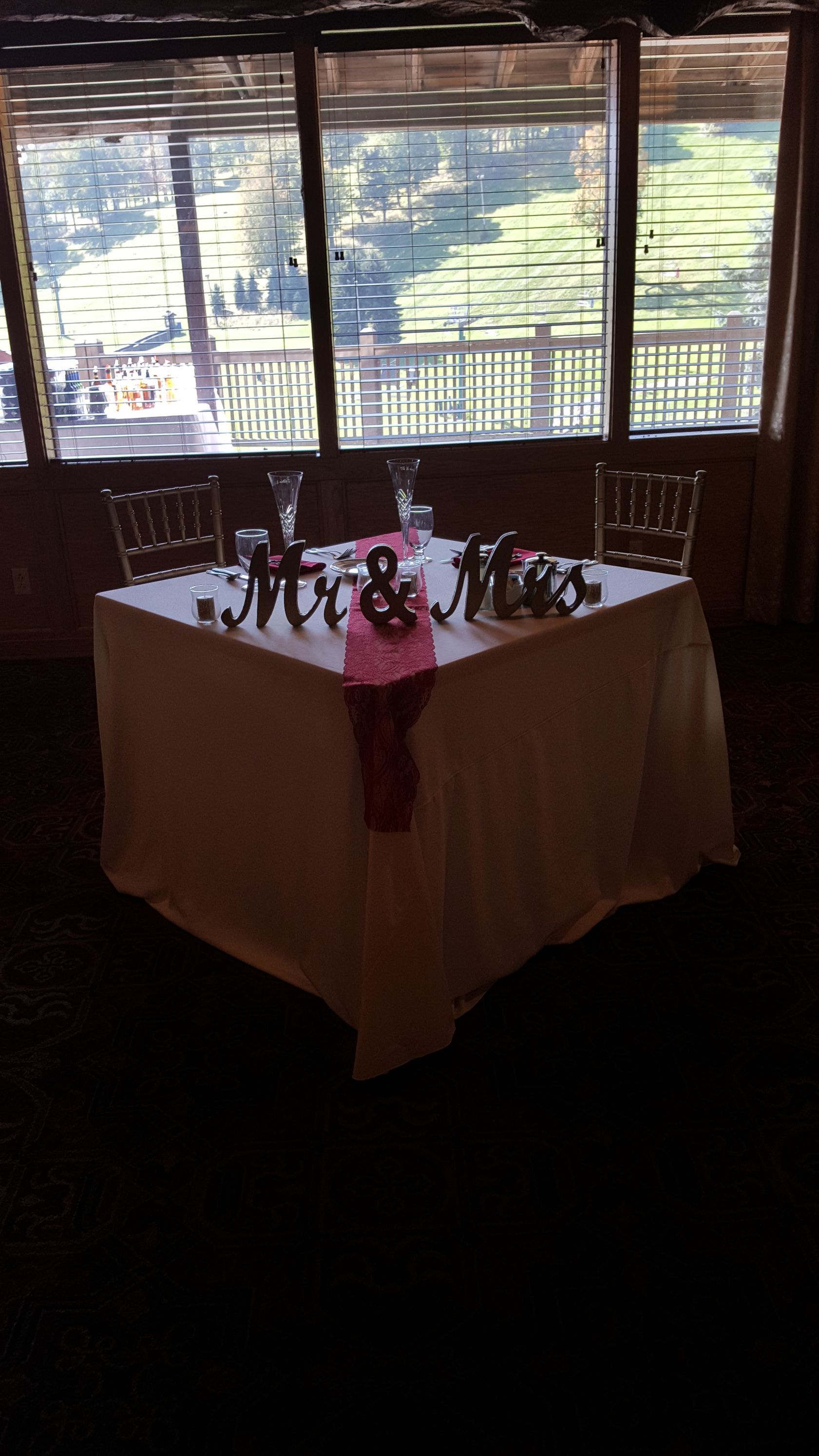 A banquet table with a white cloth and pink runner, set with plates and centerpieces by a window.
