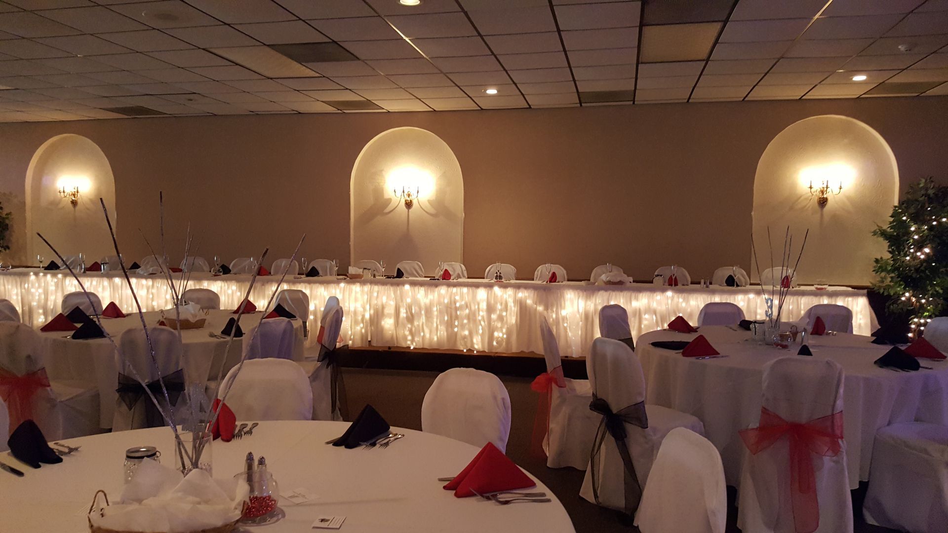 Elegant banquet hall with round tables, white chairs, red napkins, and warm wall sconces