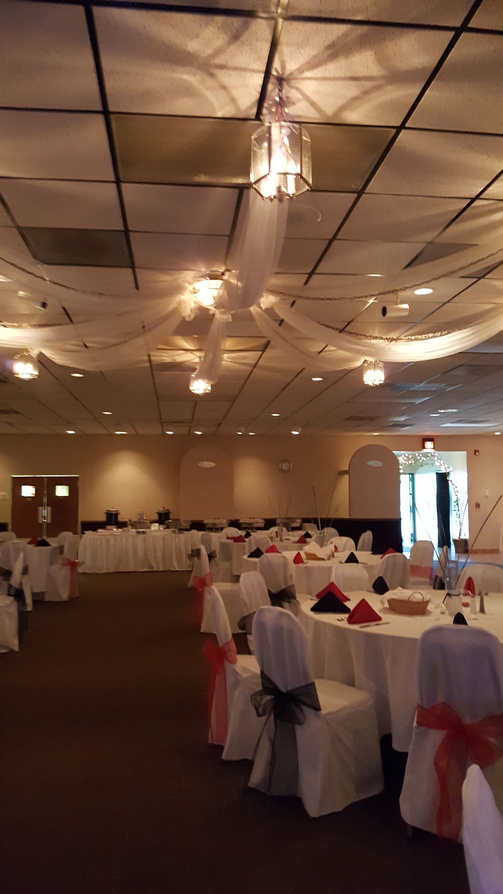 Banquet hall with white-covered chairs and tables set for an event, ceiling draped with lights.