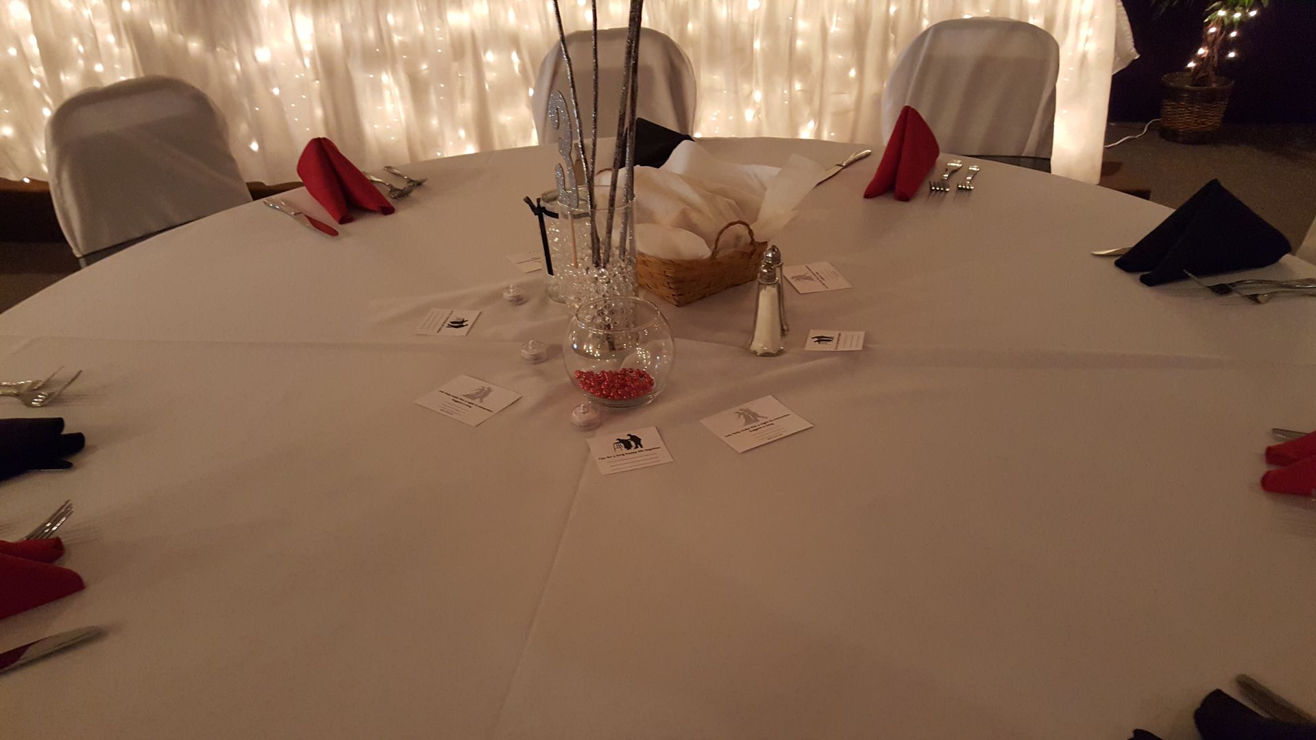 Round banquet table set with white linens, folded napkins, chairs, and soft fairy lights in the background