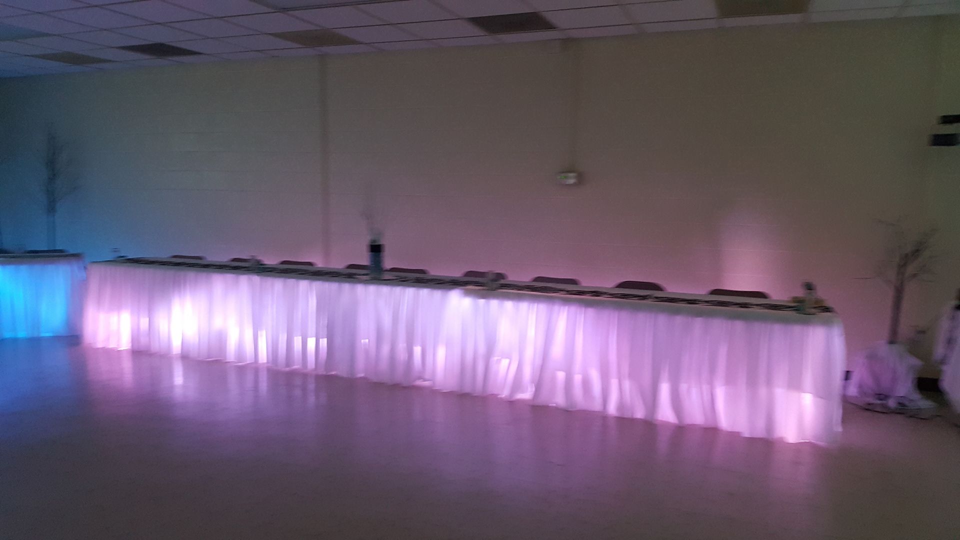 Long banquet table draped in white with purple glowing lights in a dim room
