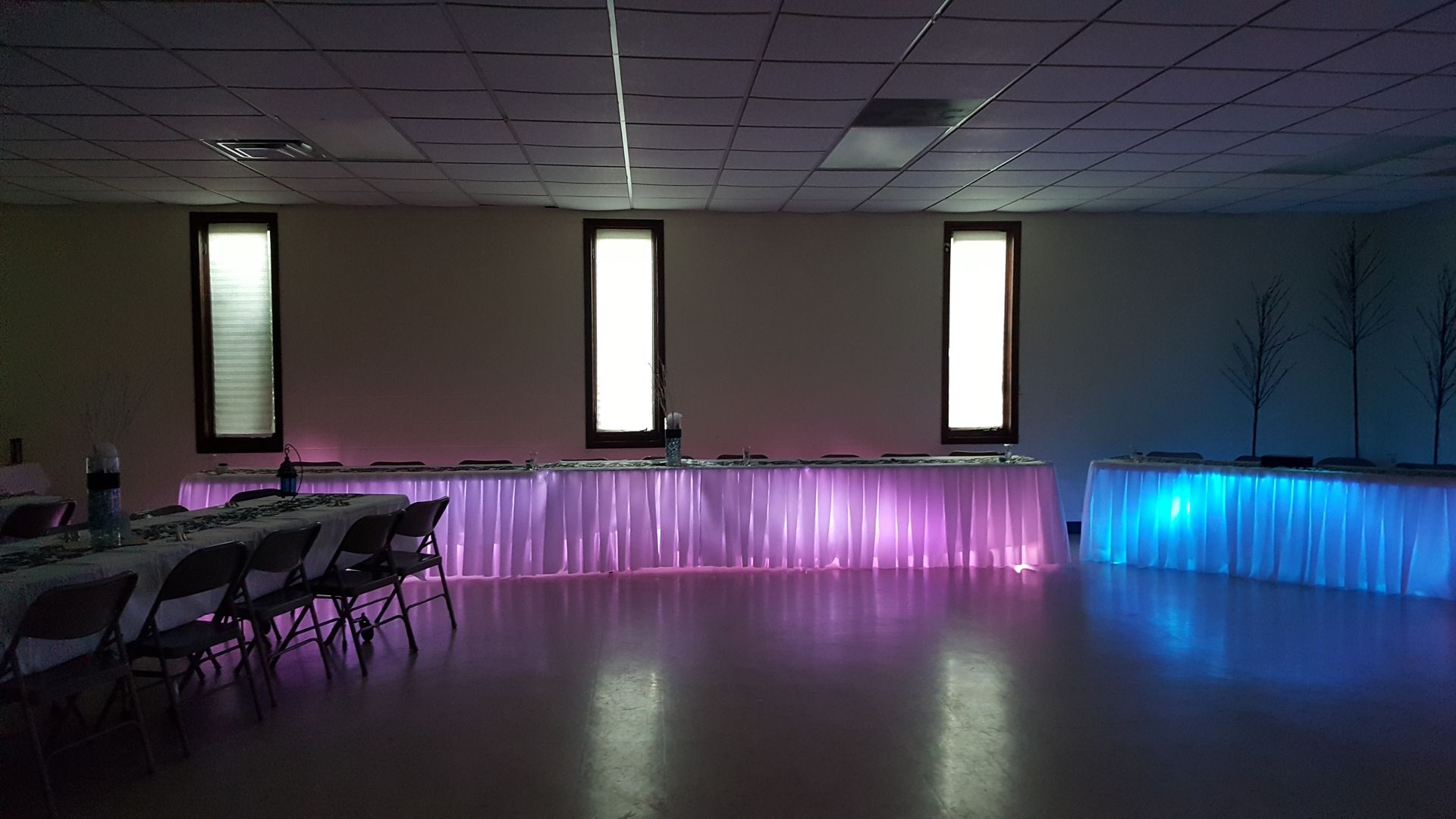 Empty event hall with folding chairs, glowing purple and blue uplighting, and three tall windows on the back wall