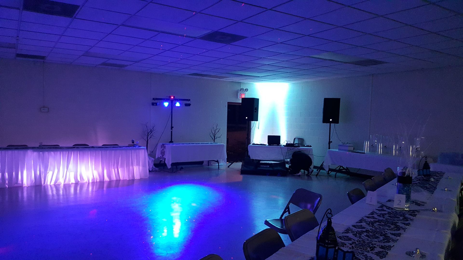 Empty event hall with blue-purple lighting, DJ setup, and tables along the right wall