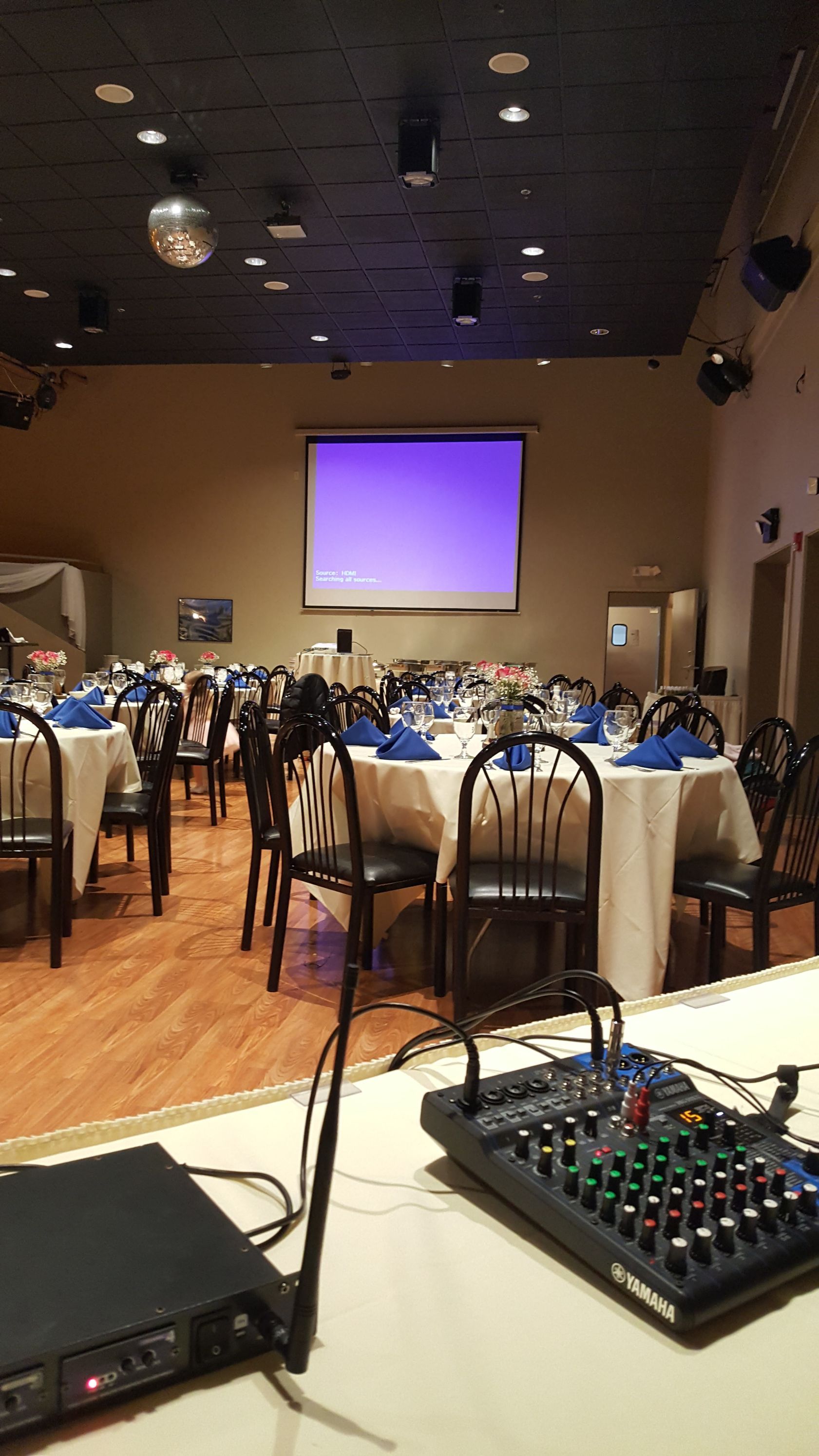Banquet hall with round tables, chairs, purple-lit projection screen, and audio mixer in the foreground