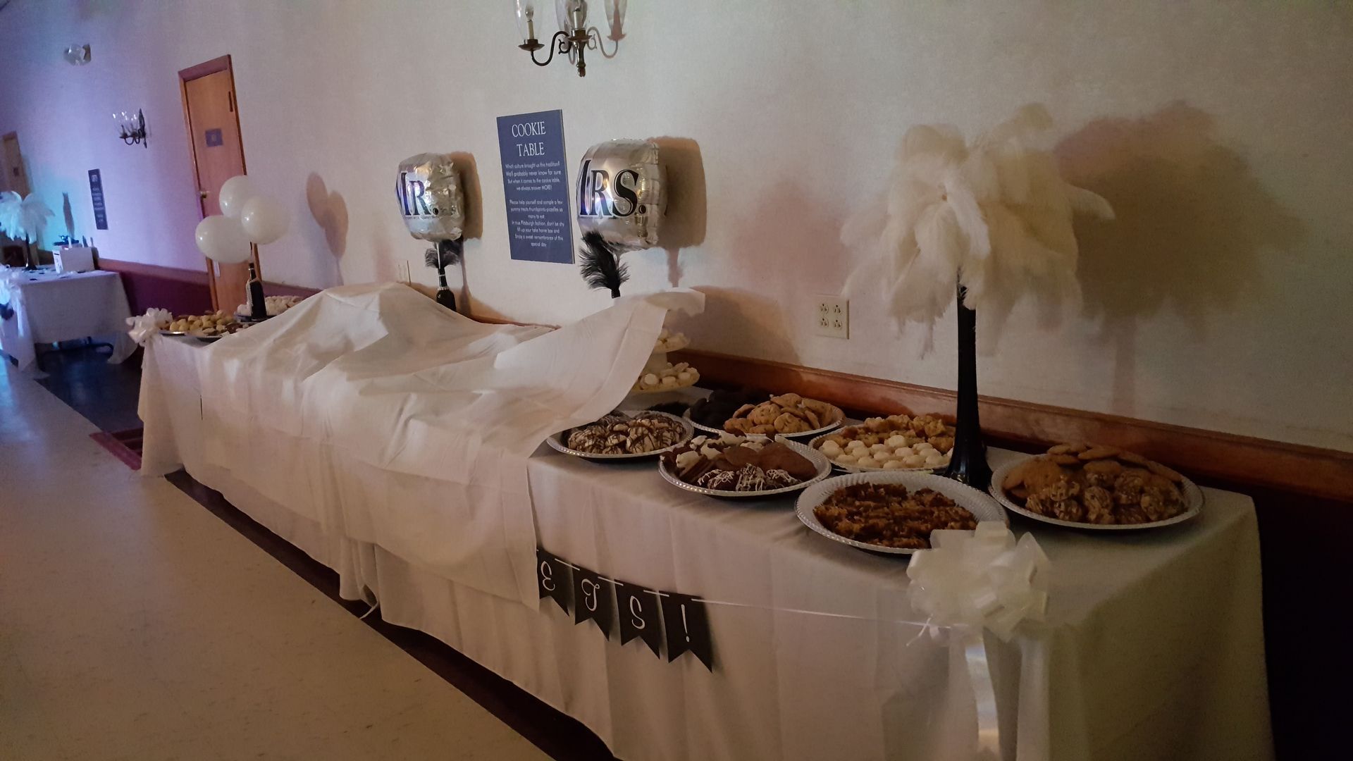 Long buffet table with white tablecloth, chafing dishes, and serving platters in a banquet hall.