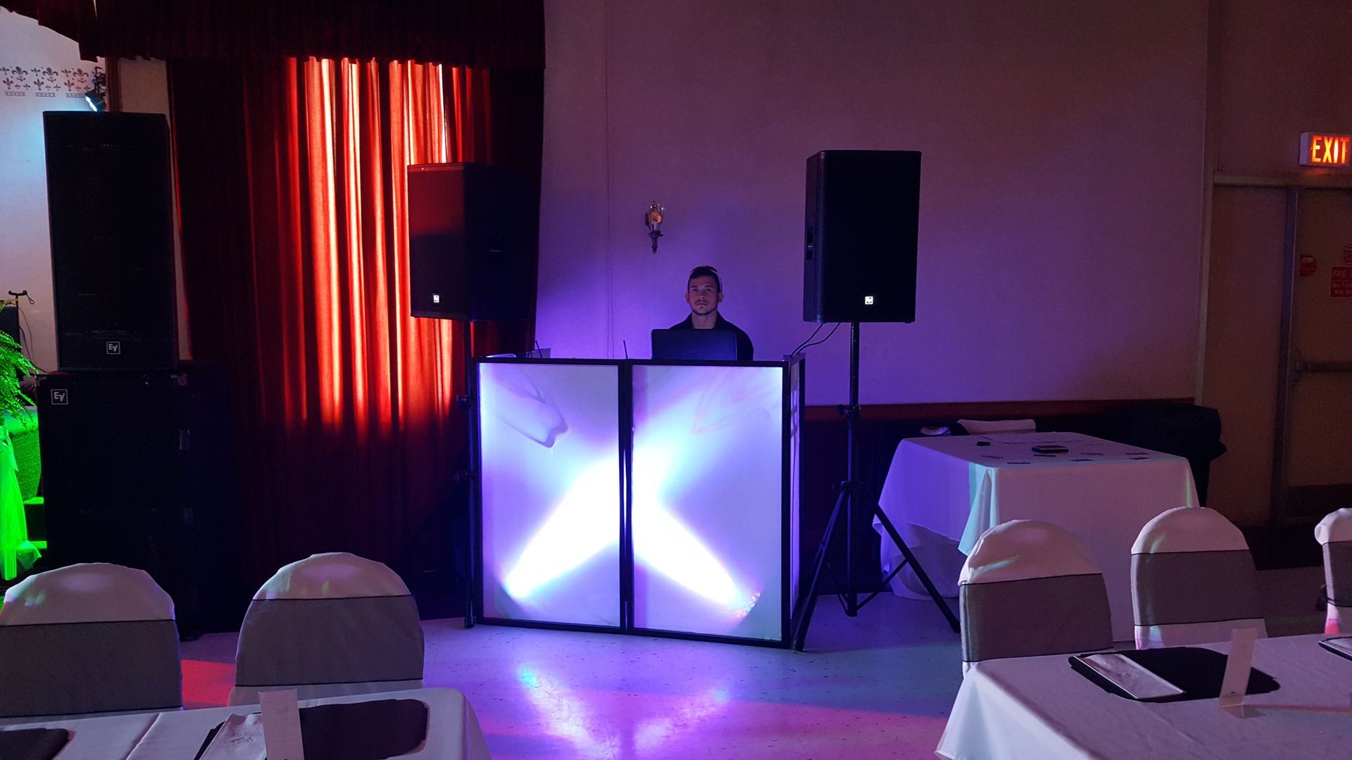 DJ behind illuminated booth in a dim event hall with tables, chairs, speakers, and red stage curtain