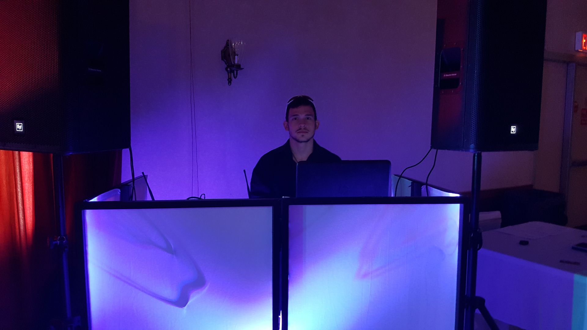 DJ standing behind a lit booth with purple and blue lights in a dark room
