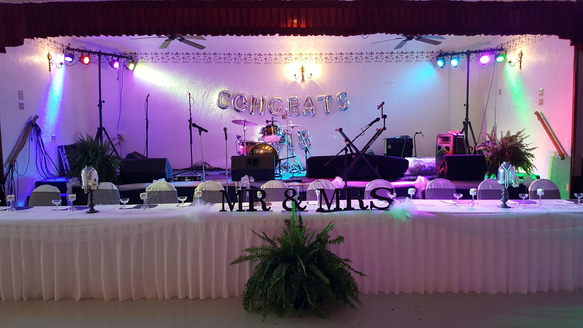 Decorated wedding stage with white drapes, lights, floral arrangements, and “Mr & Mrs” sign